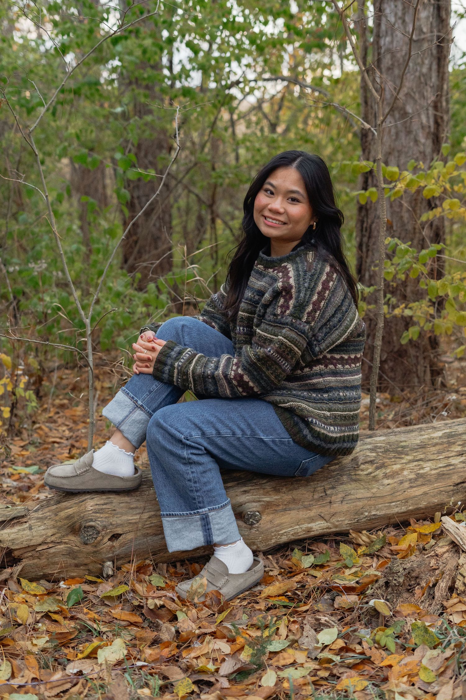 Someone sits on a fallen log in autumn woods wearing a patterned sweater and jeans surrounded by fallen leaves.