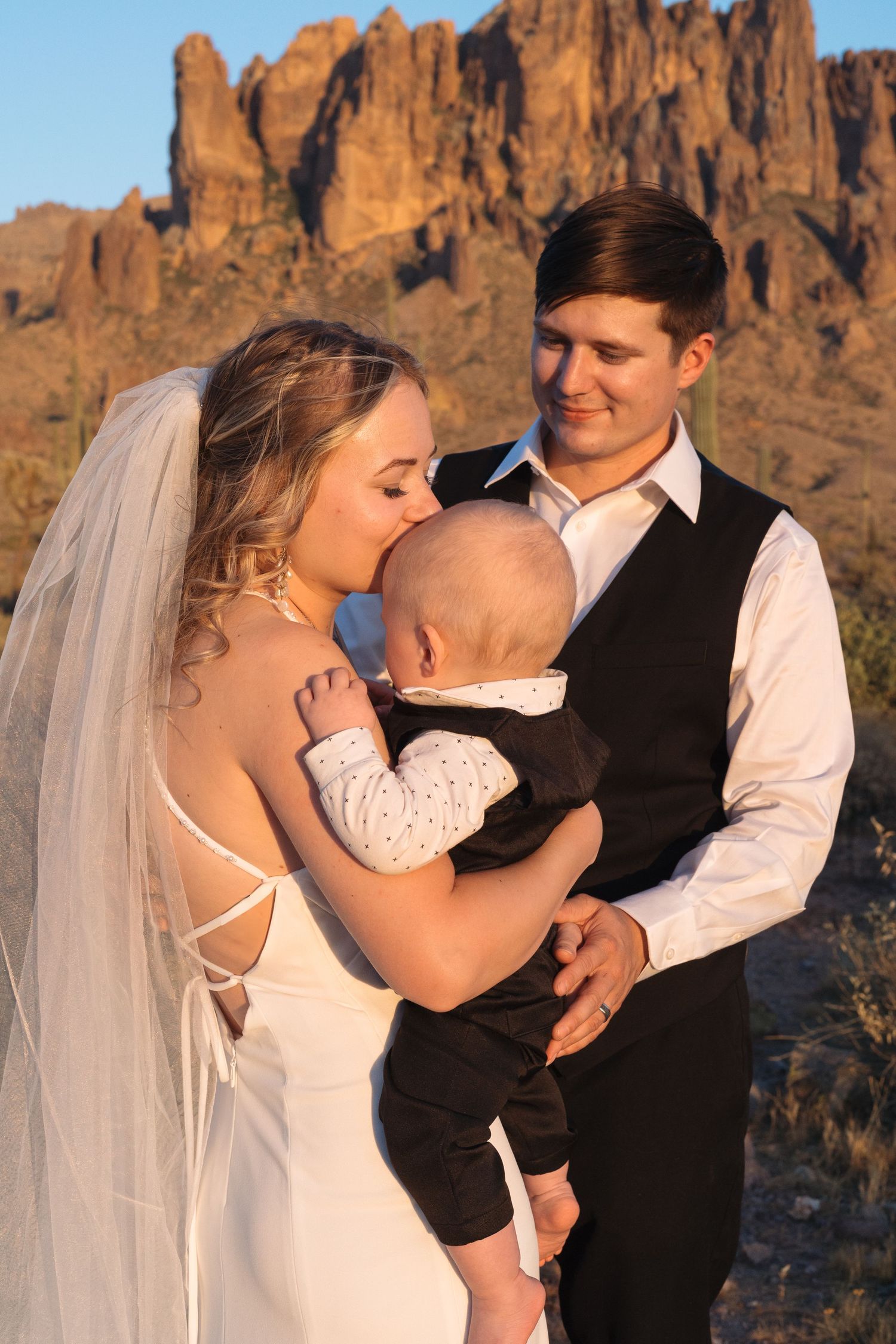 Bride and groom holding baby at desert wedding with Superstition Mountains backdrop in Arizona golden hour light.