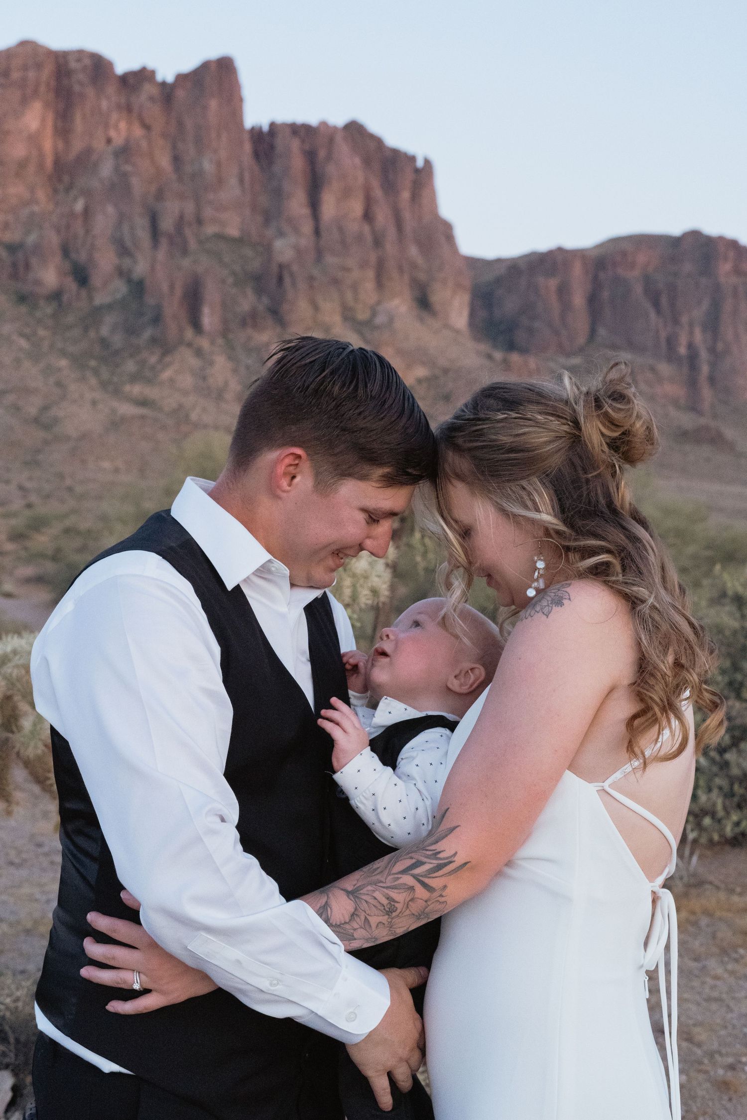 Wedding couple in desert landscape sharing tender moment with baby, red rock mountains in background.