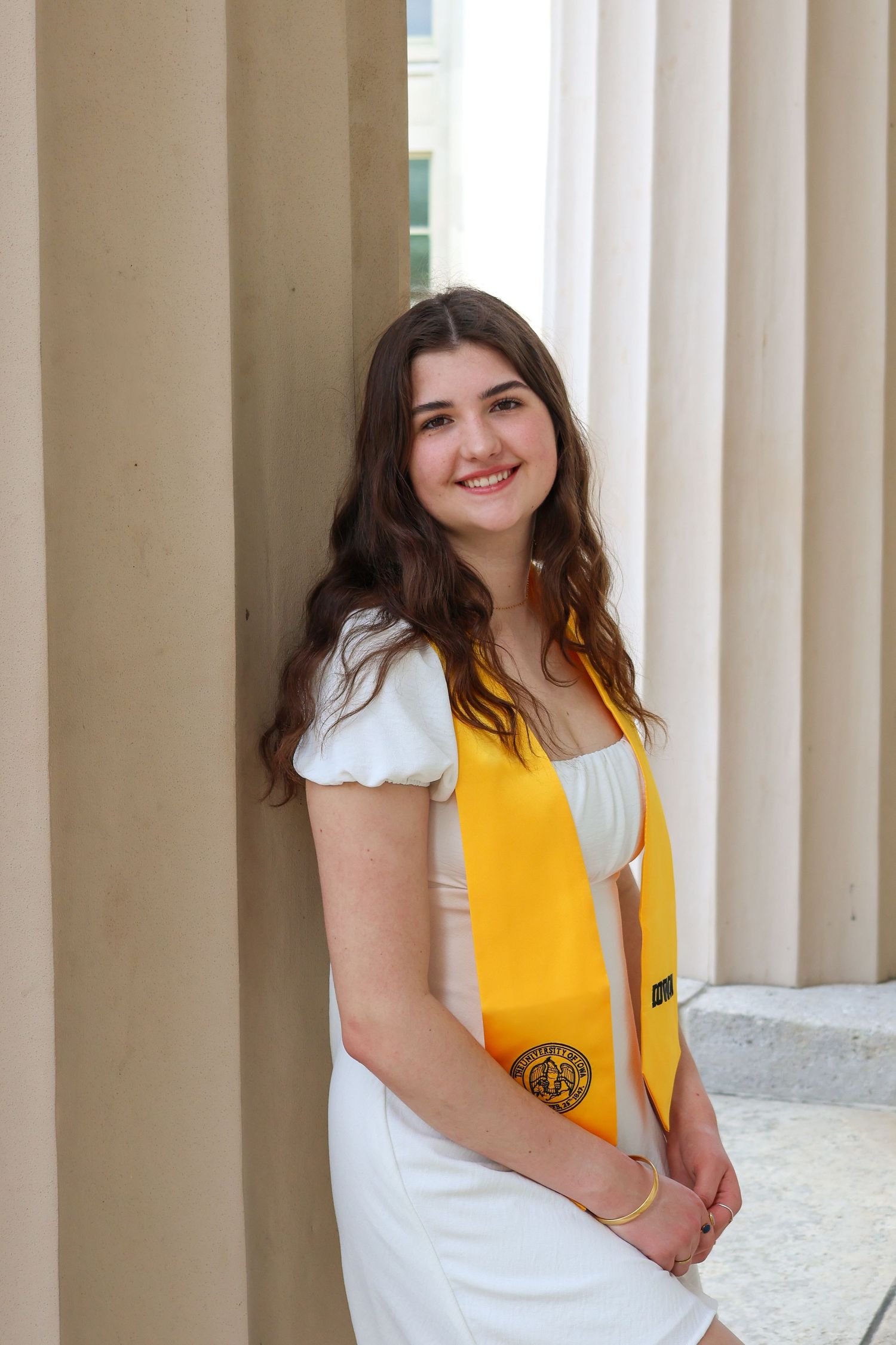 A graduate in a white dress and yellow stole poses between classical columns.
