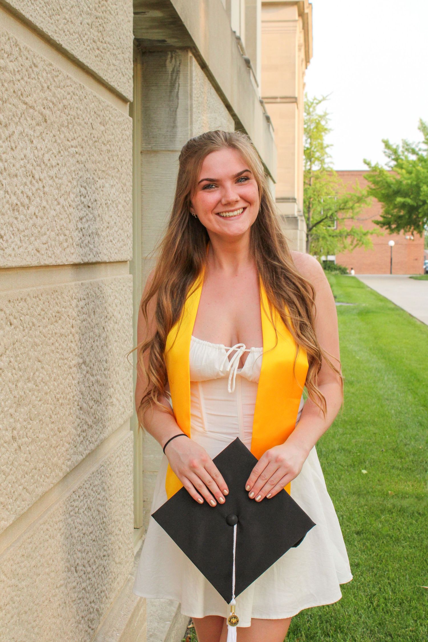 Graduate in yellow stole holds black graduation cap in front of campus building.