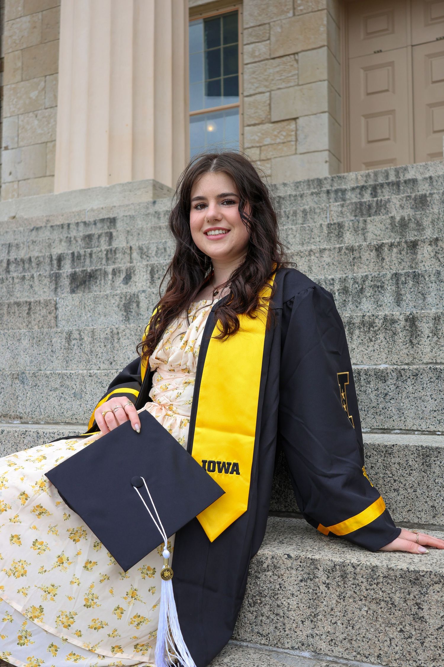Graduate in academic regalia poses on stone steps with University of Iowa banners visible.
