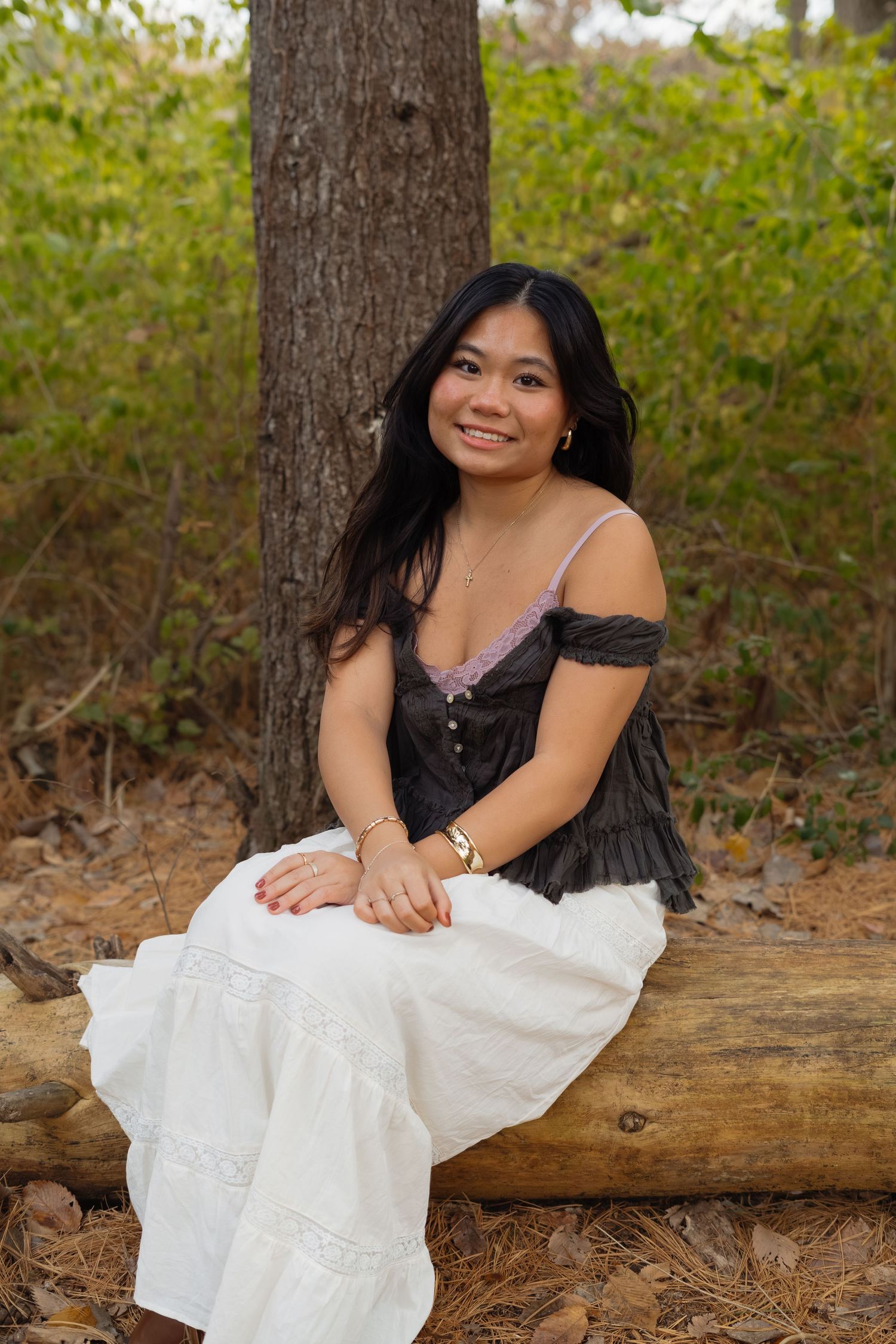 A person in a black and white dress sits on a fallen log in a wooded outdoor setting.