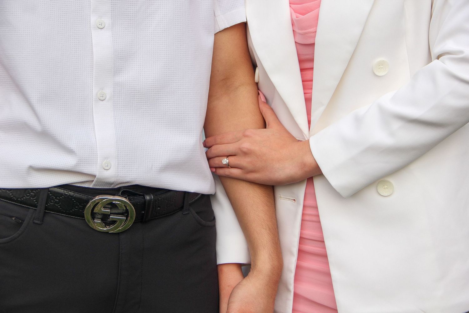 A couple in formal attire stands close together, with one wearing a white blazer and the other in a white shirt.