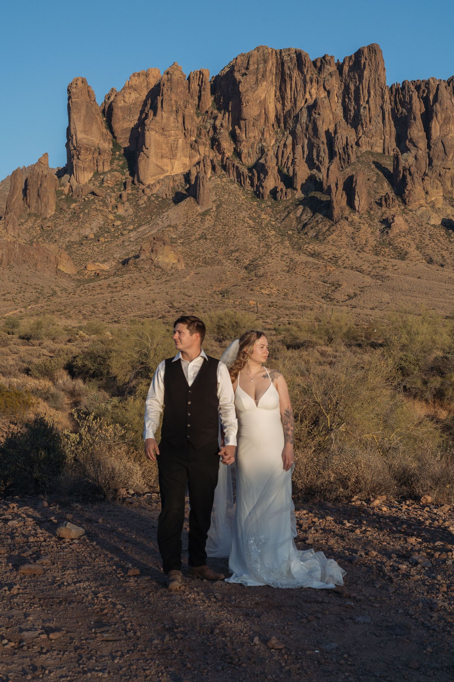 Bride and groom hold hands in desert landscape with Superstition Mountains at golden hour in Arizona.