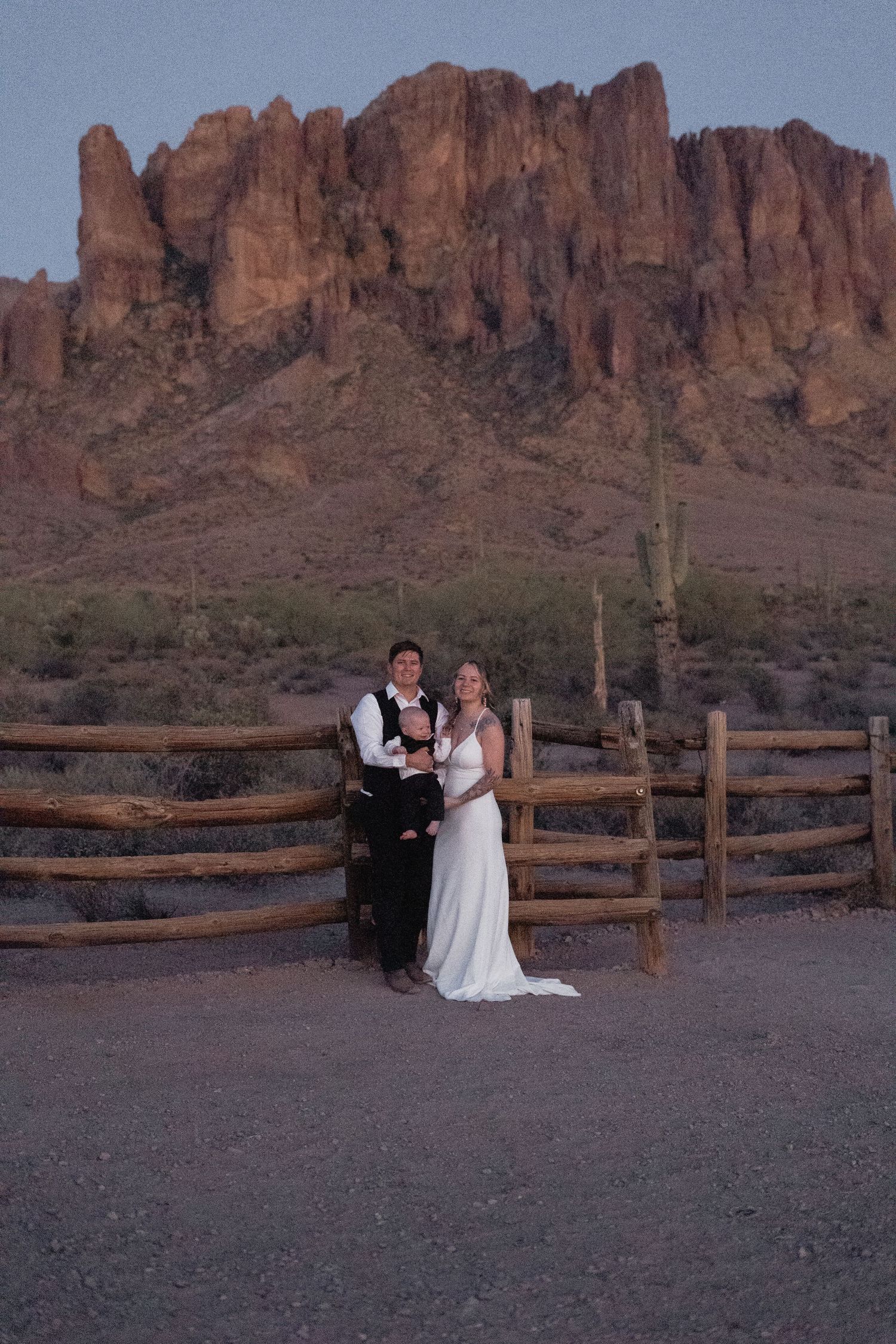 Bride and groom pose by rustic fence with Superstition Mountains at sunset in Arizona desert wedding photo.