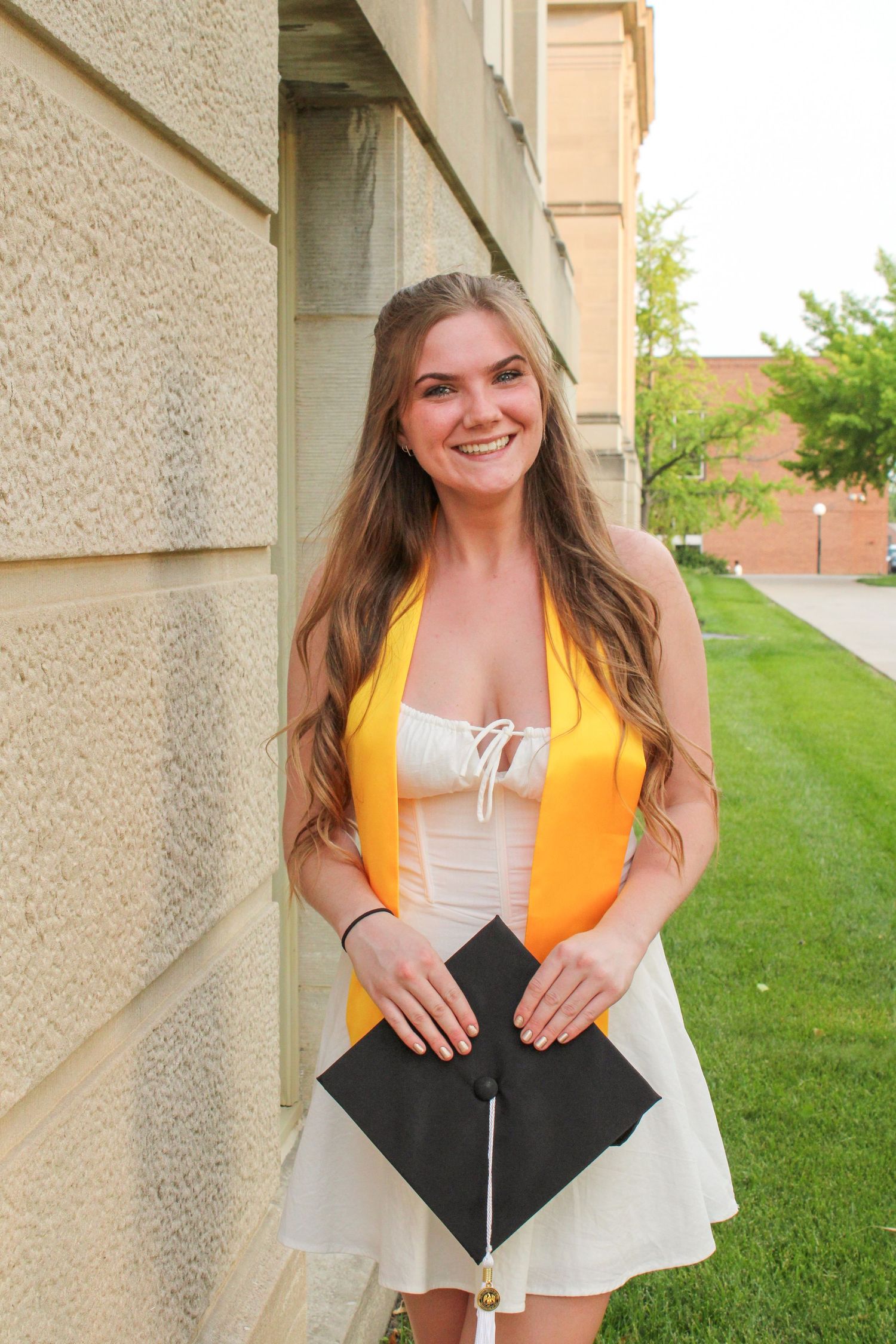 A series of graduation photos showing someone in a gold stole and white dress holding their cap against a building exterior.