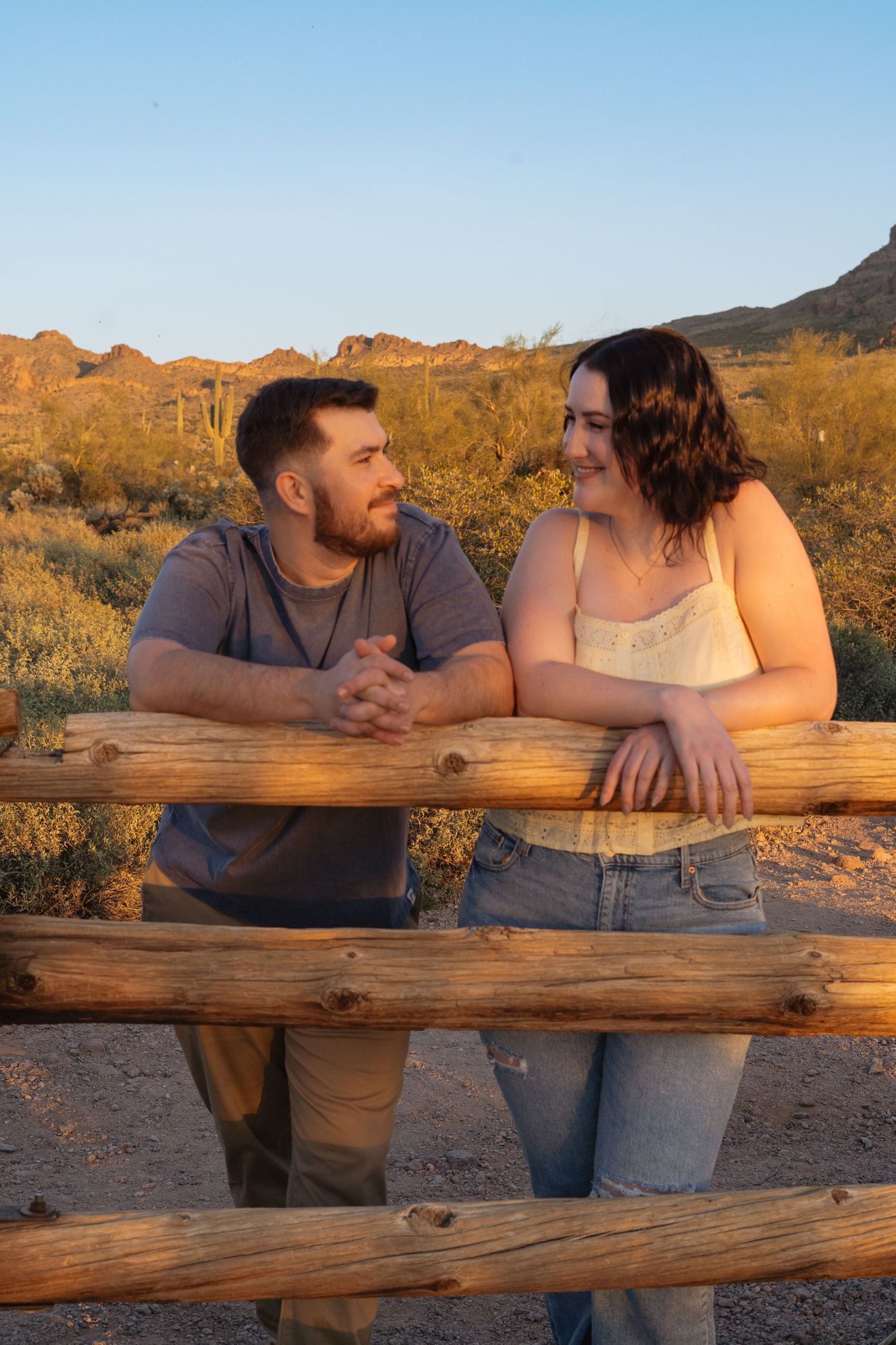 A couple leans on a wooden fence at sunset in a desert landscape with golden light and desert flora in the background.