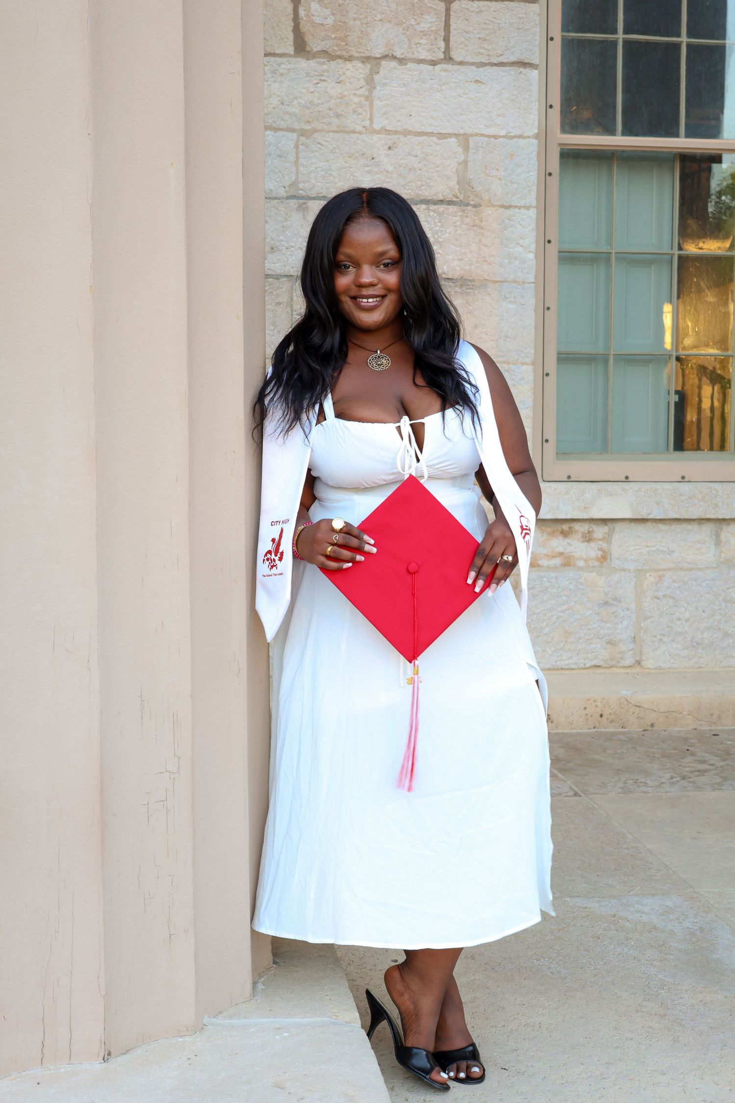 Graduate in white dress poses with red graduation cap in front of stone columns.