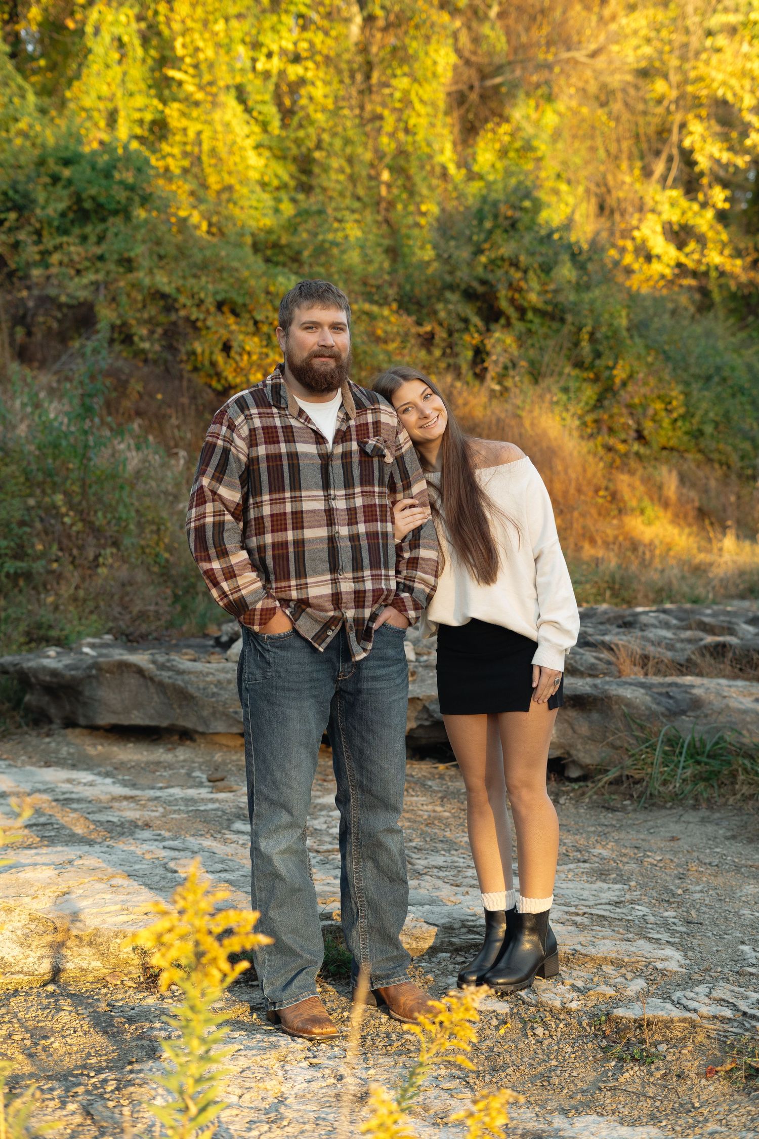A couple stands together on rocky terrain surrounded by golden autumn foliage during a sunset photoshoot.