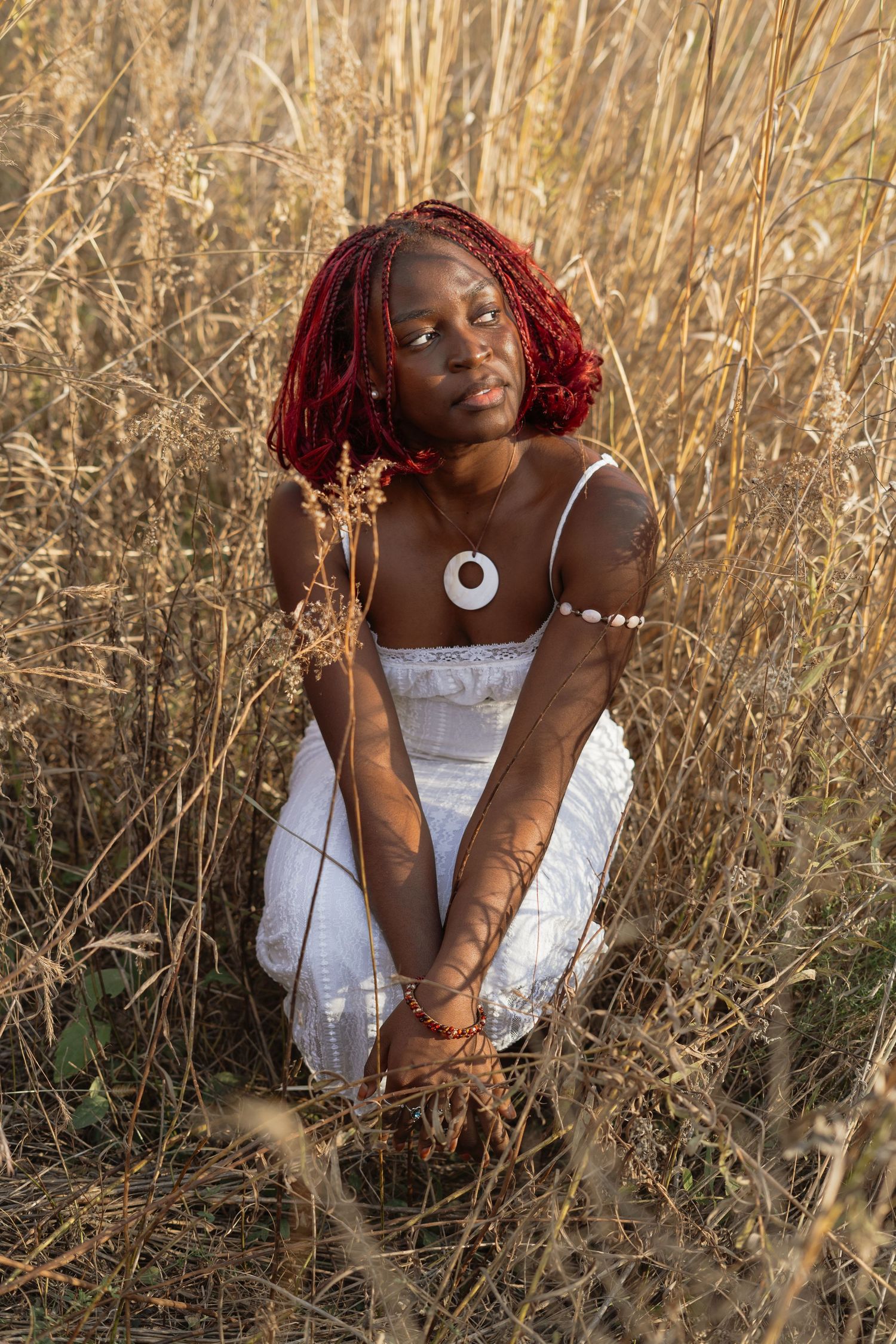 Person in white dress and red headwrap sits thoughtfully among tall golden grass in warm sunlight.