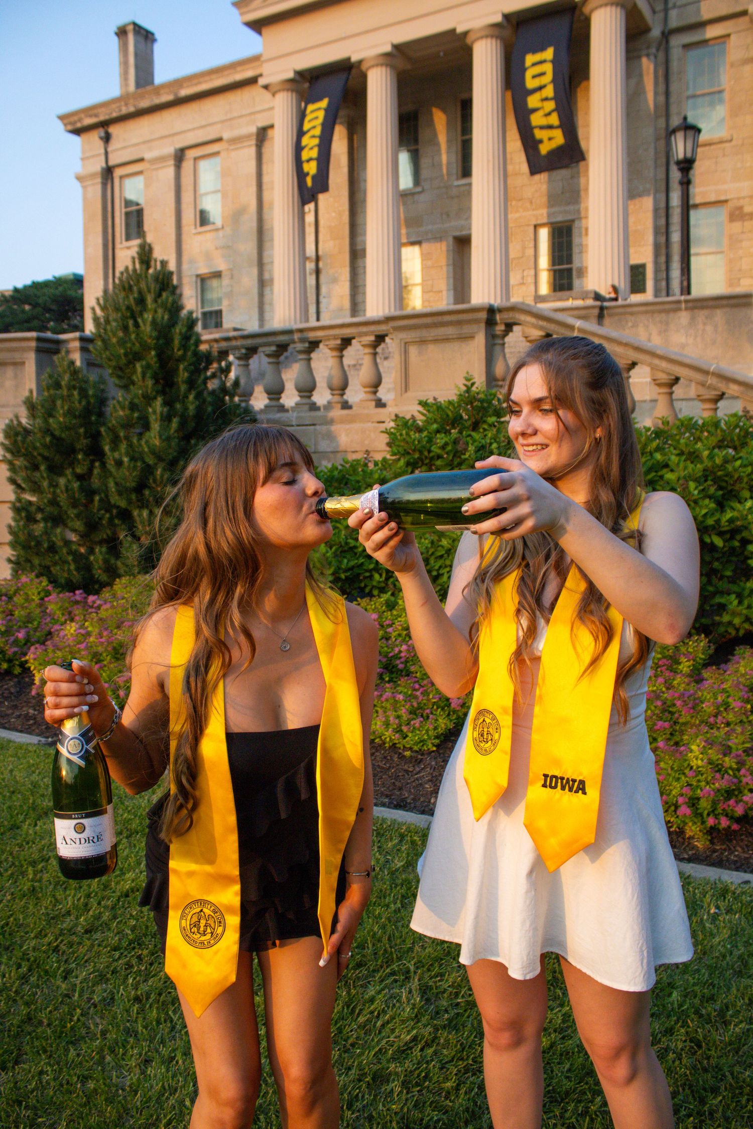 Two graduates in yellow stoles celebrate with champagne outside historic university building.