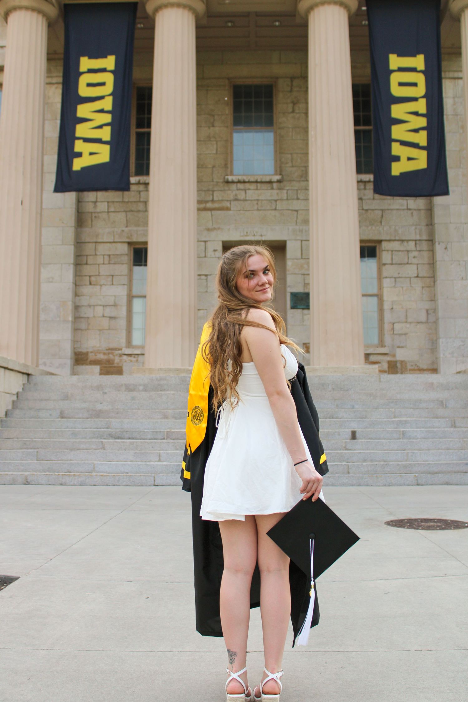 Graduate poses on stone steps of university building with yellow and black banners.