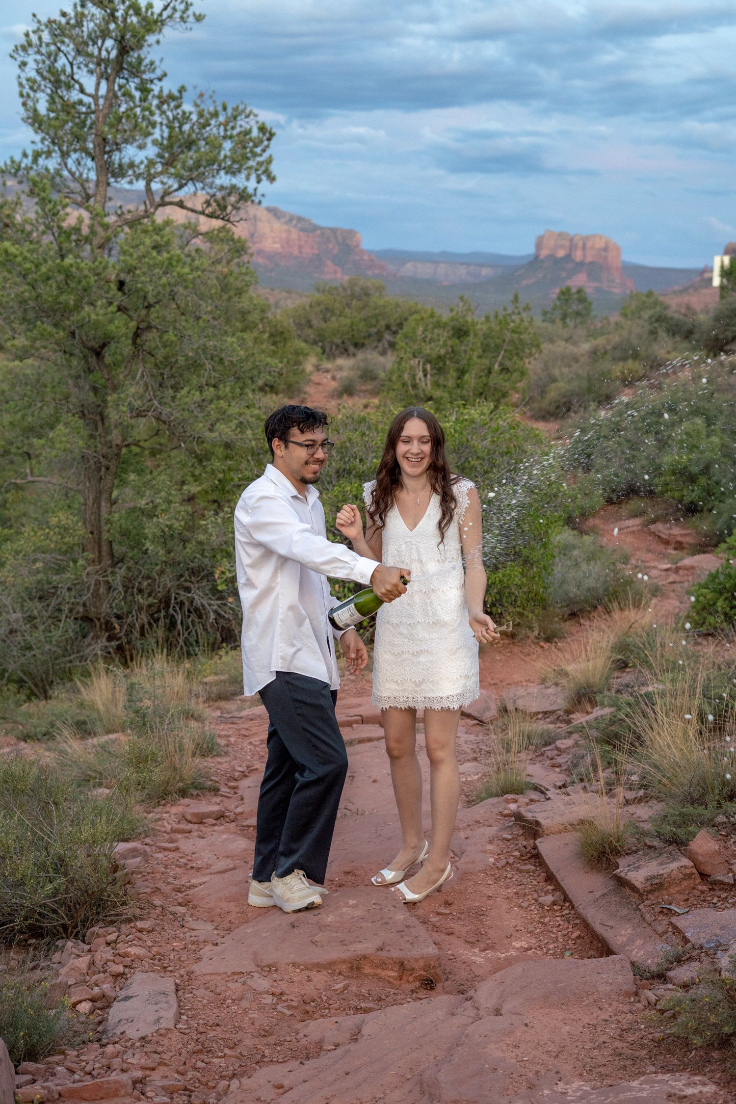 Couple celebrating with champagne on red rock trail in Sedona, Arizona, with stunning butte formations in background.