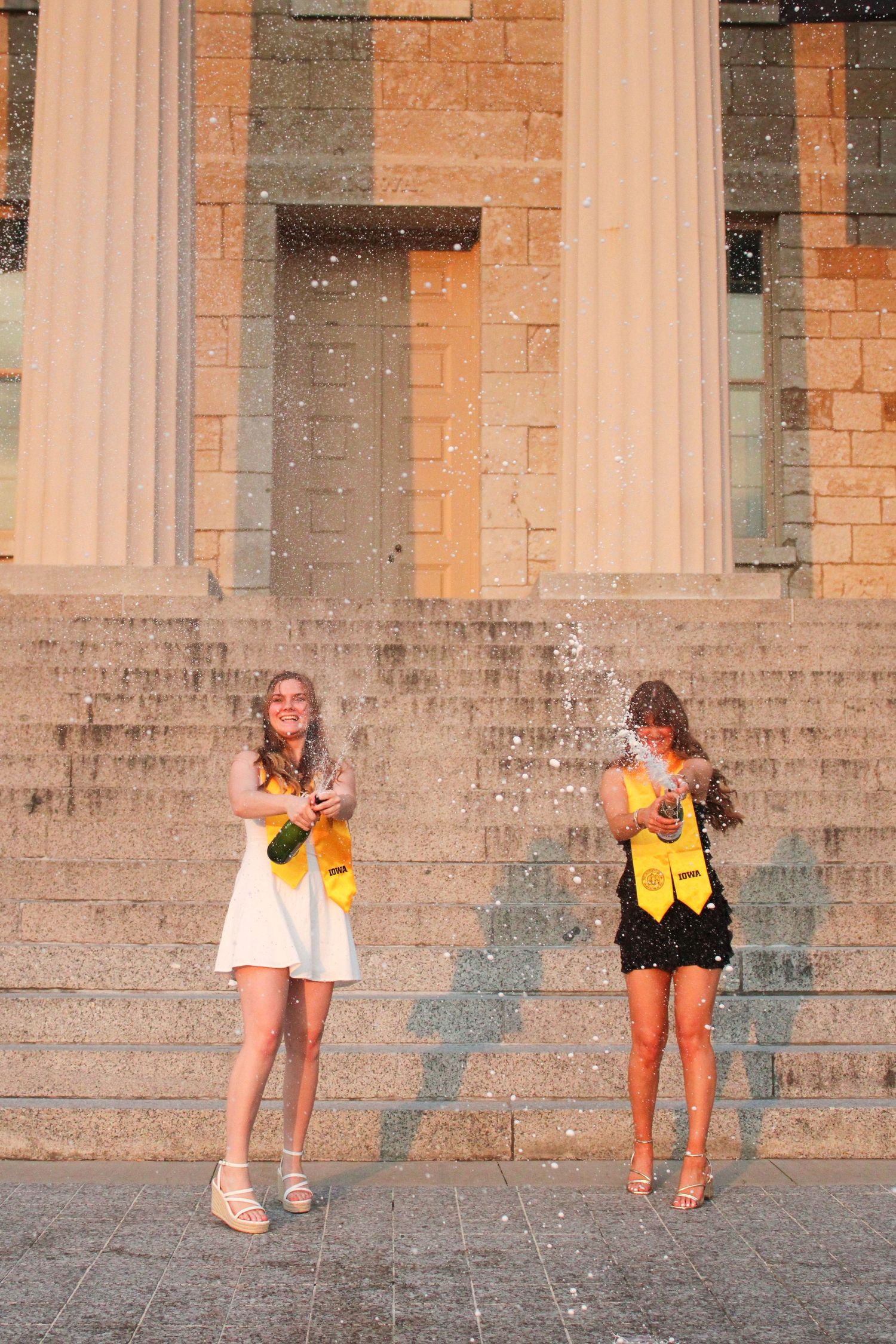 Two people playfully jump in front of a stone building with columns.