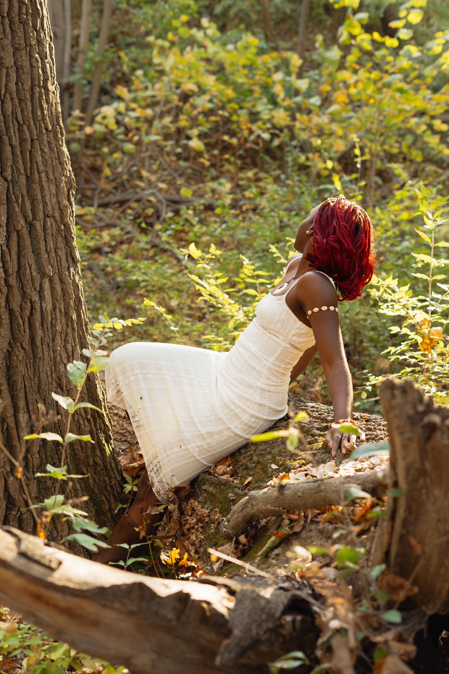 A figure in a white dress lies peacefully among fallen leaves and tree trunks in a sunlit autumn forest setting.