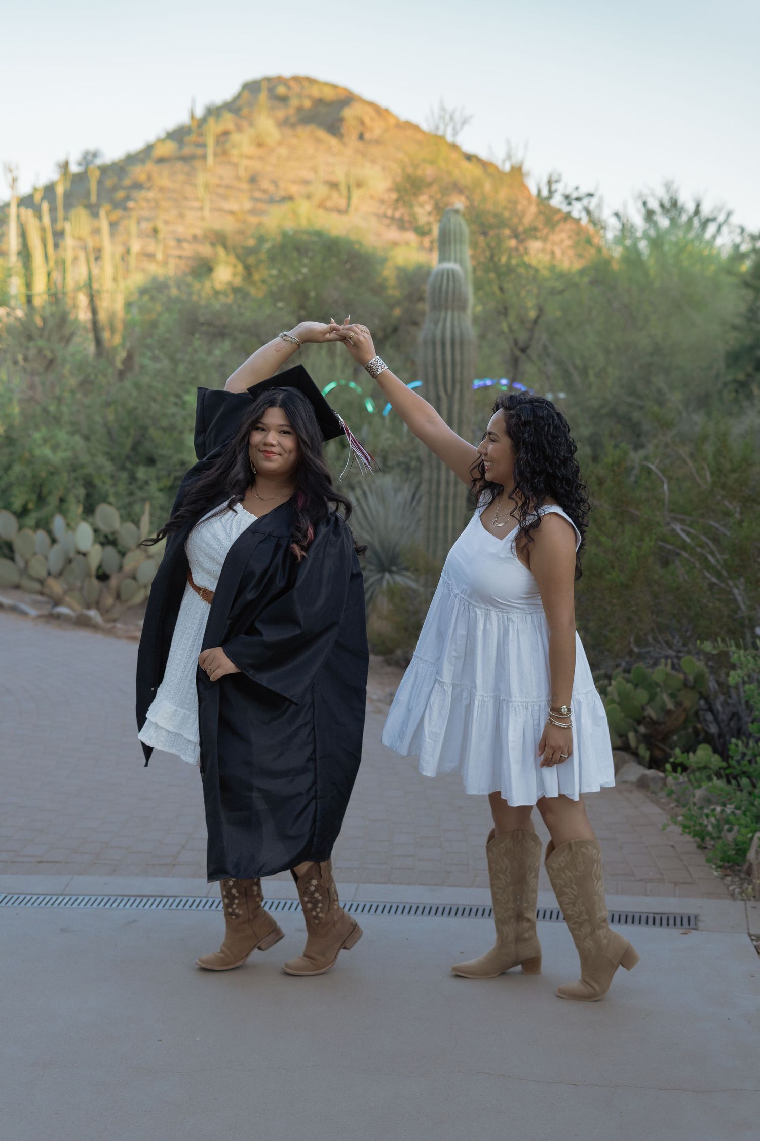 Two women celebrate graduation outdoors in Arizona desert, one in cap and gown, with saguaro cacti.