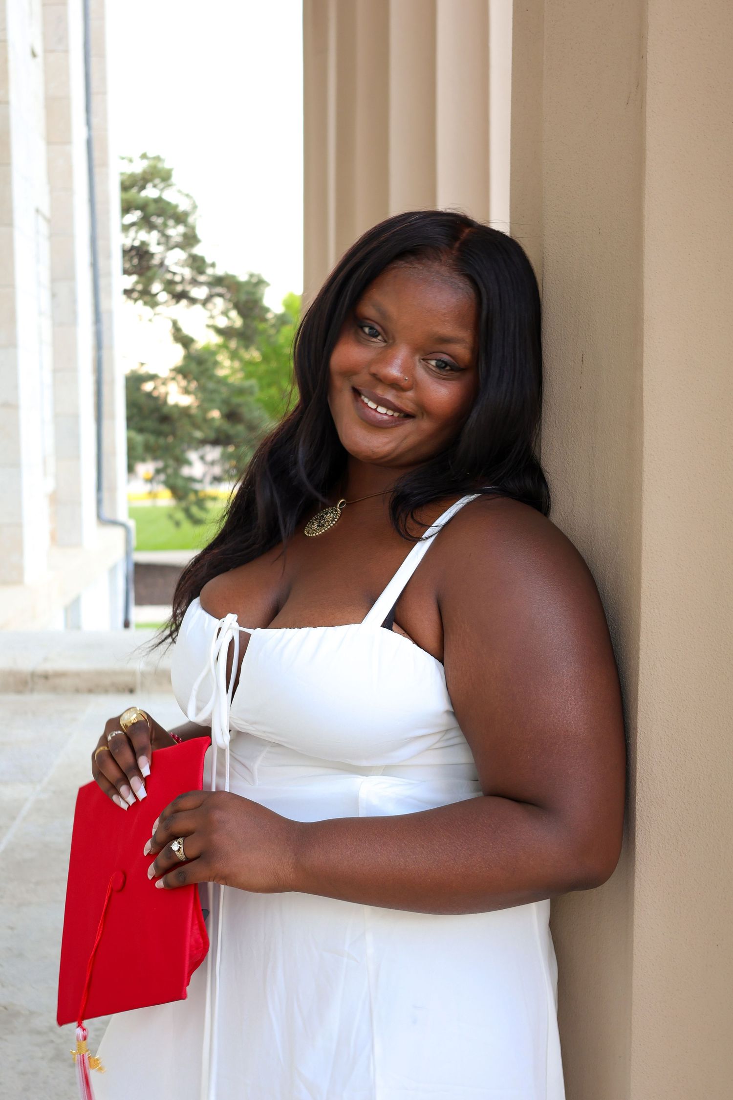 Graduate in white dress smiles while holding red folder against stone column backdrop.