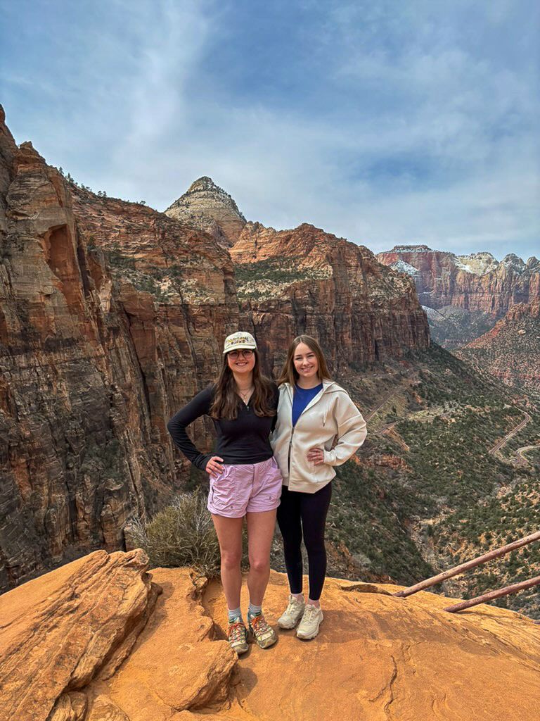 Two hikers stand on red rock formations overlooking Zion National Park's dramatic canyon landscape.