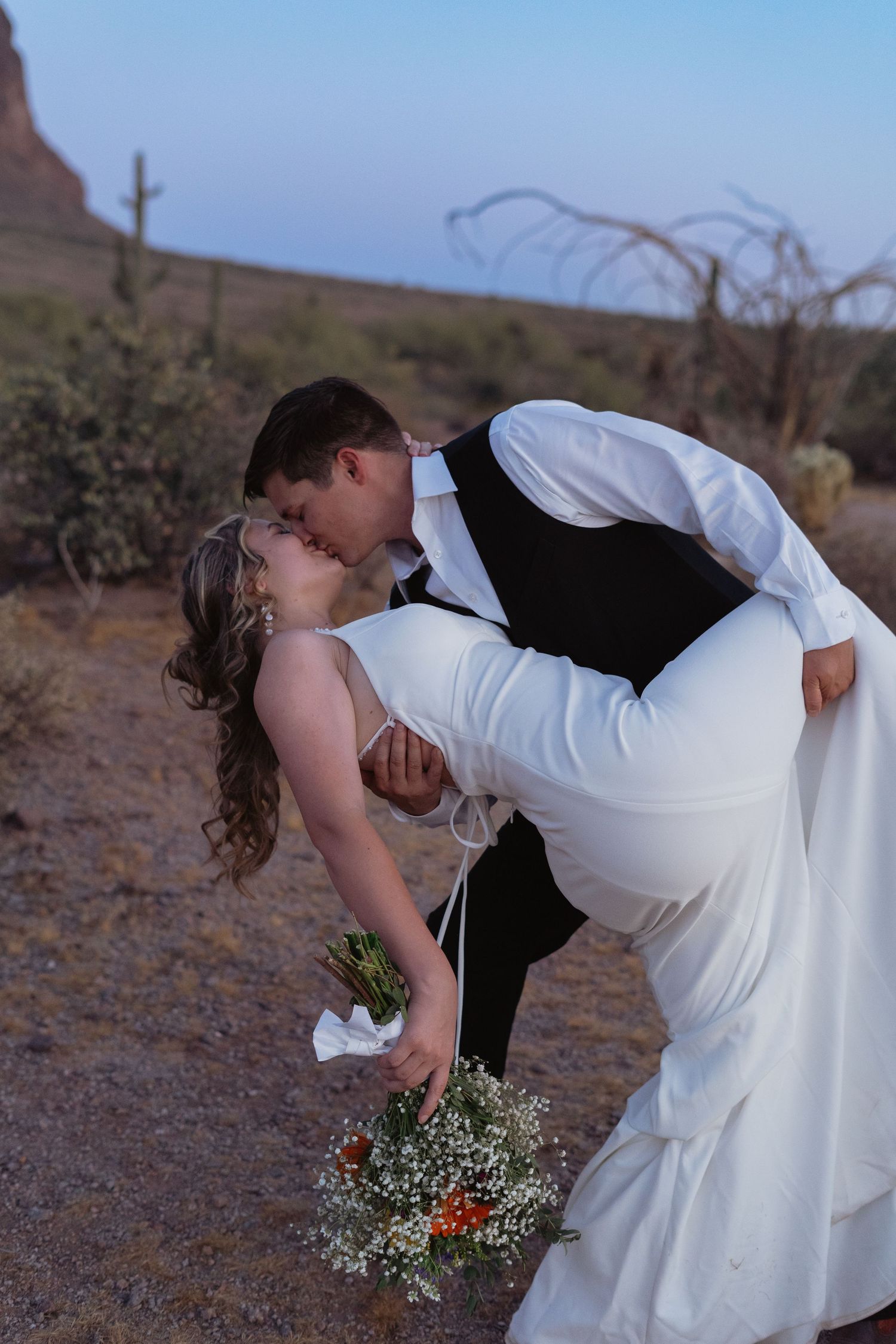 Groom dips bride for a romantic kiss in desert landscape, bride holding wildflower bouquet at sunset.