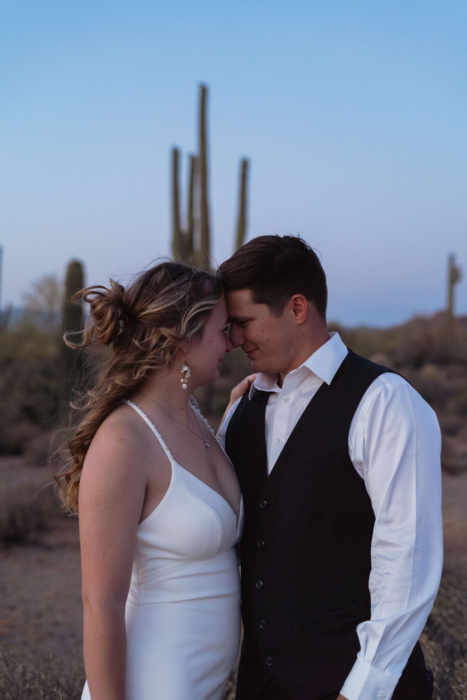 A bride and groom share an intimate moment in the Arizona desert at dusk, with saguaro cacti in the background.