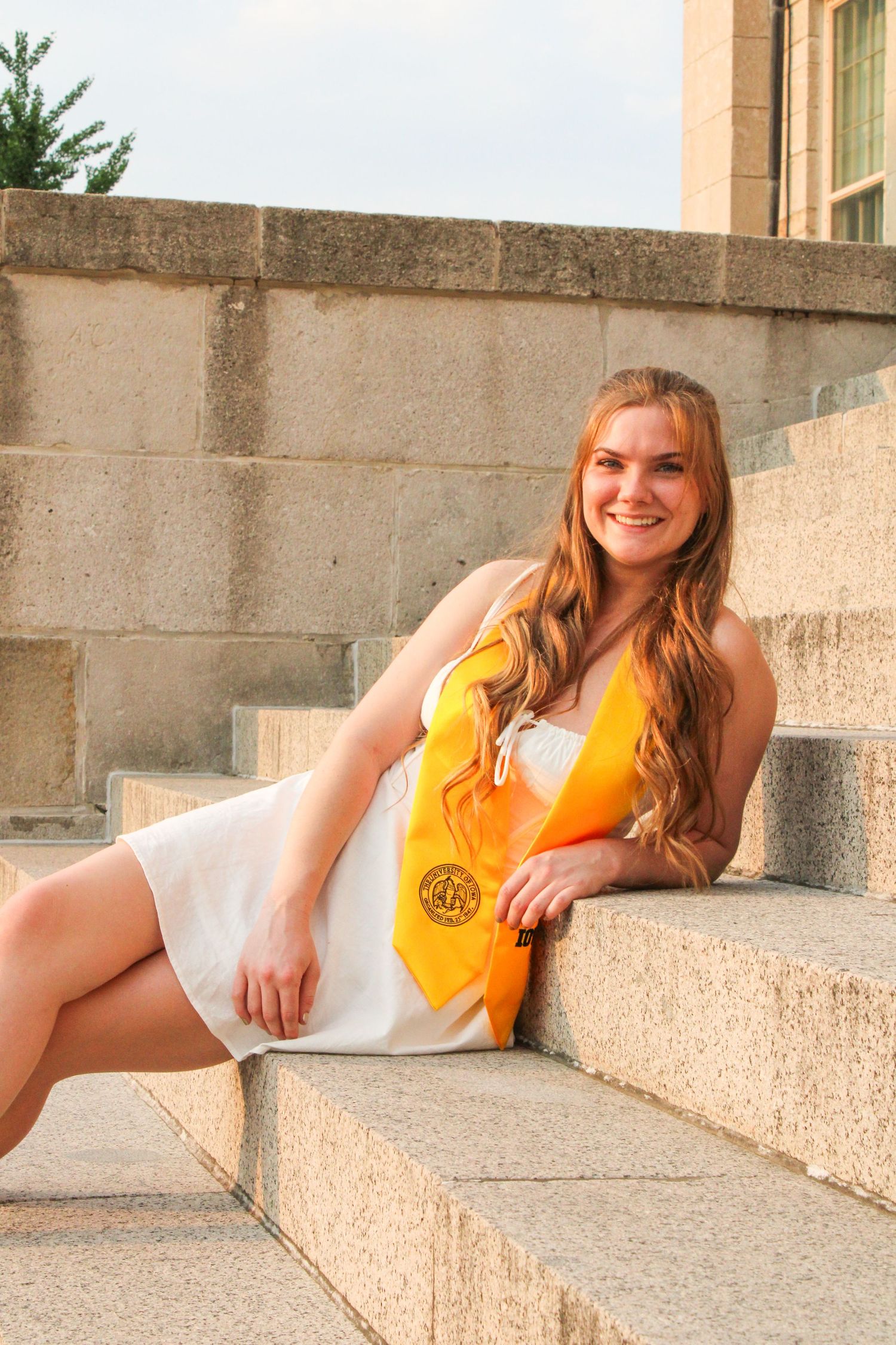 A graduate in white dress and yellow sash poses on concrete steps during golden hour.