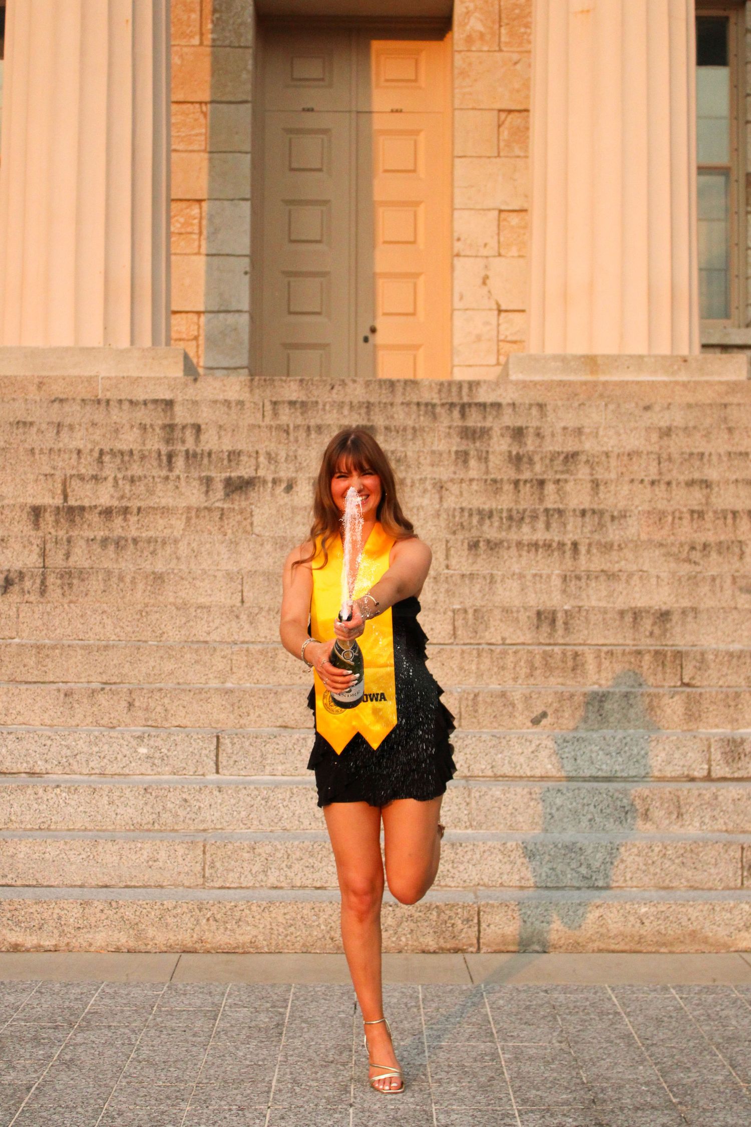 A graduate poses on stone steps wearing a black dress and yellow sash in warm sunlight.