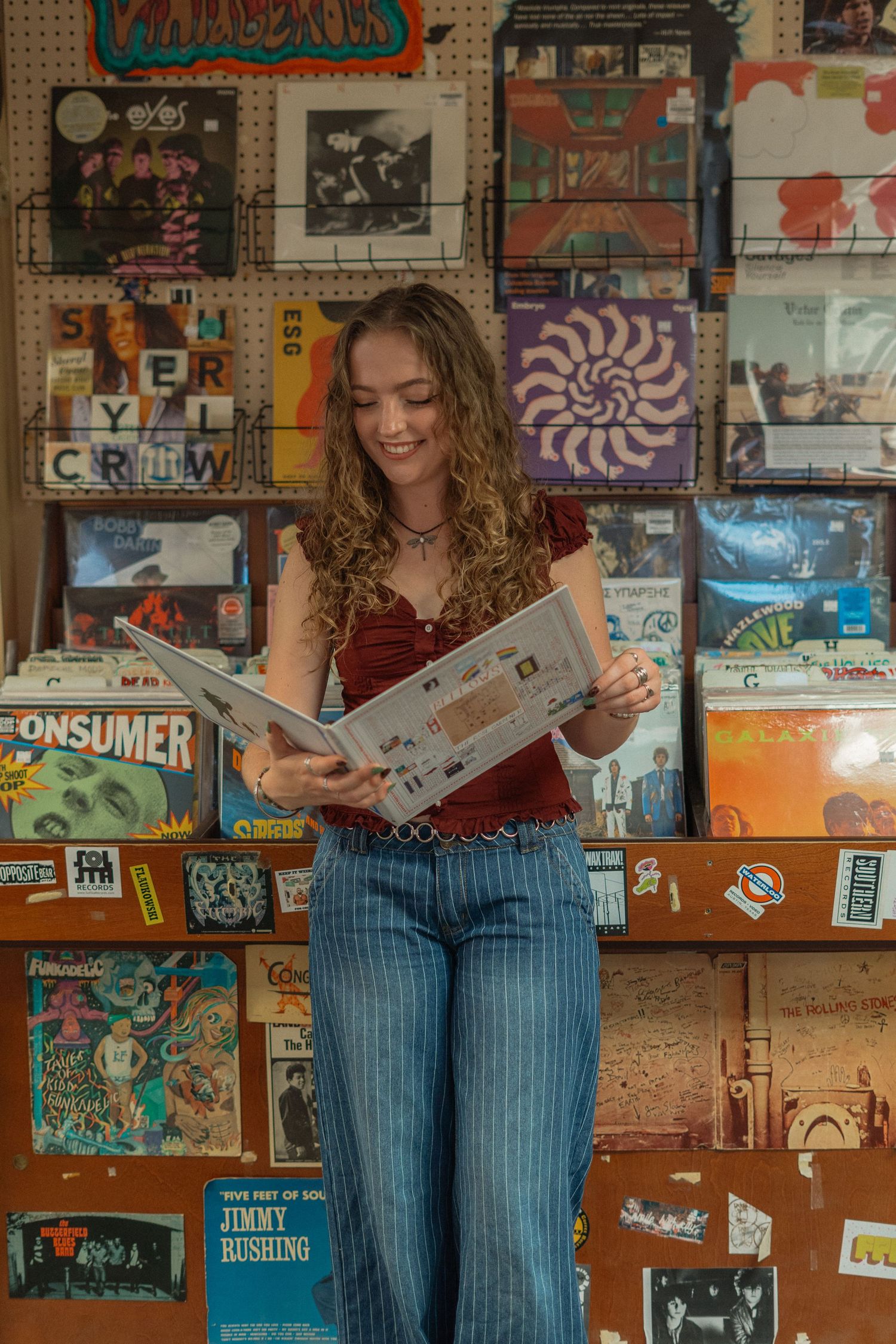 Person in striped jeans sits reading on a colorful wall covered with posters and artwork in a casual setting.