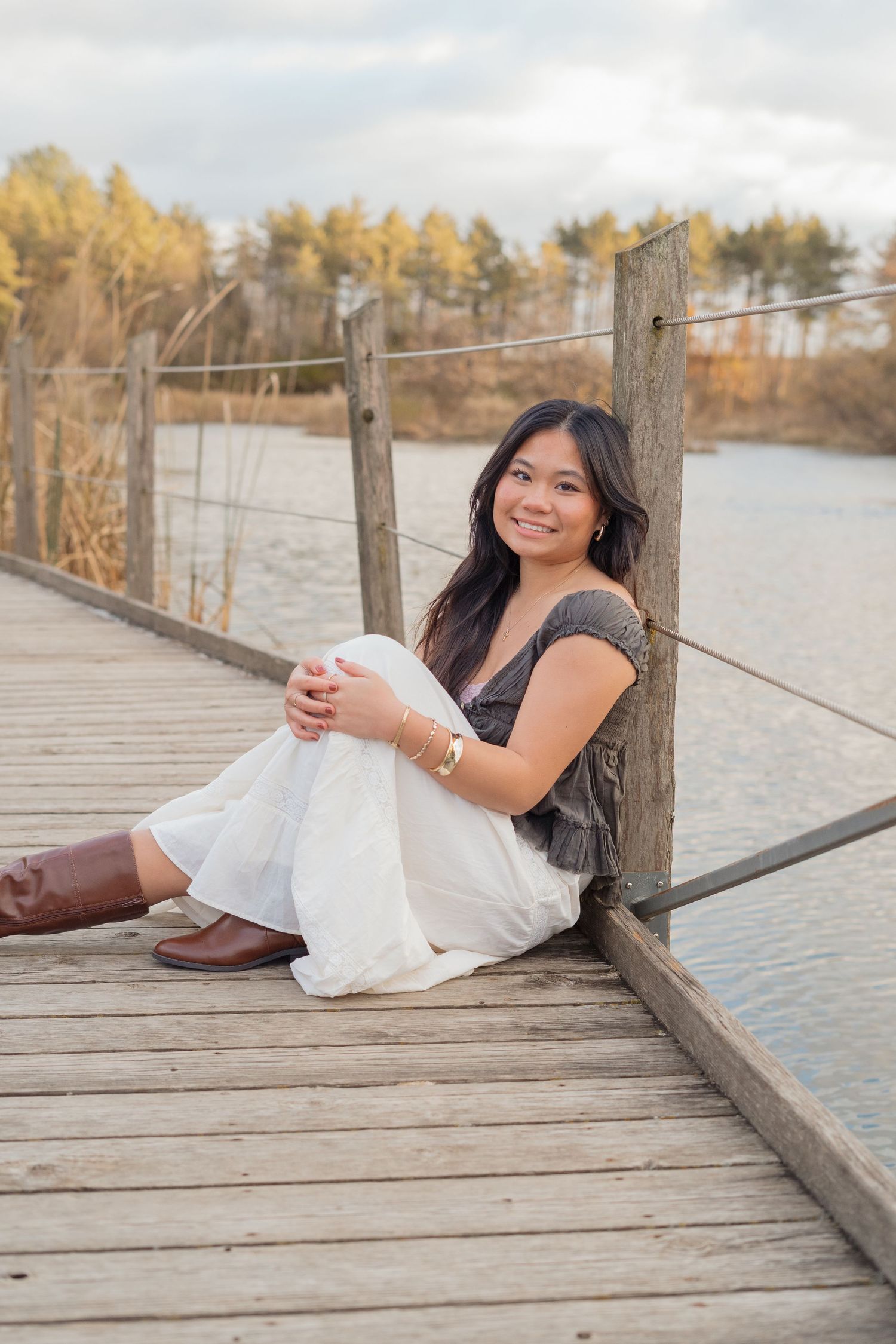 A person in a white dress reclines on a wooden dock by a lake during golden hour with autumn trees in the background.