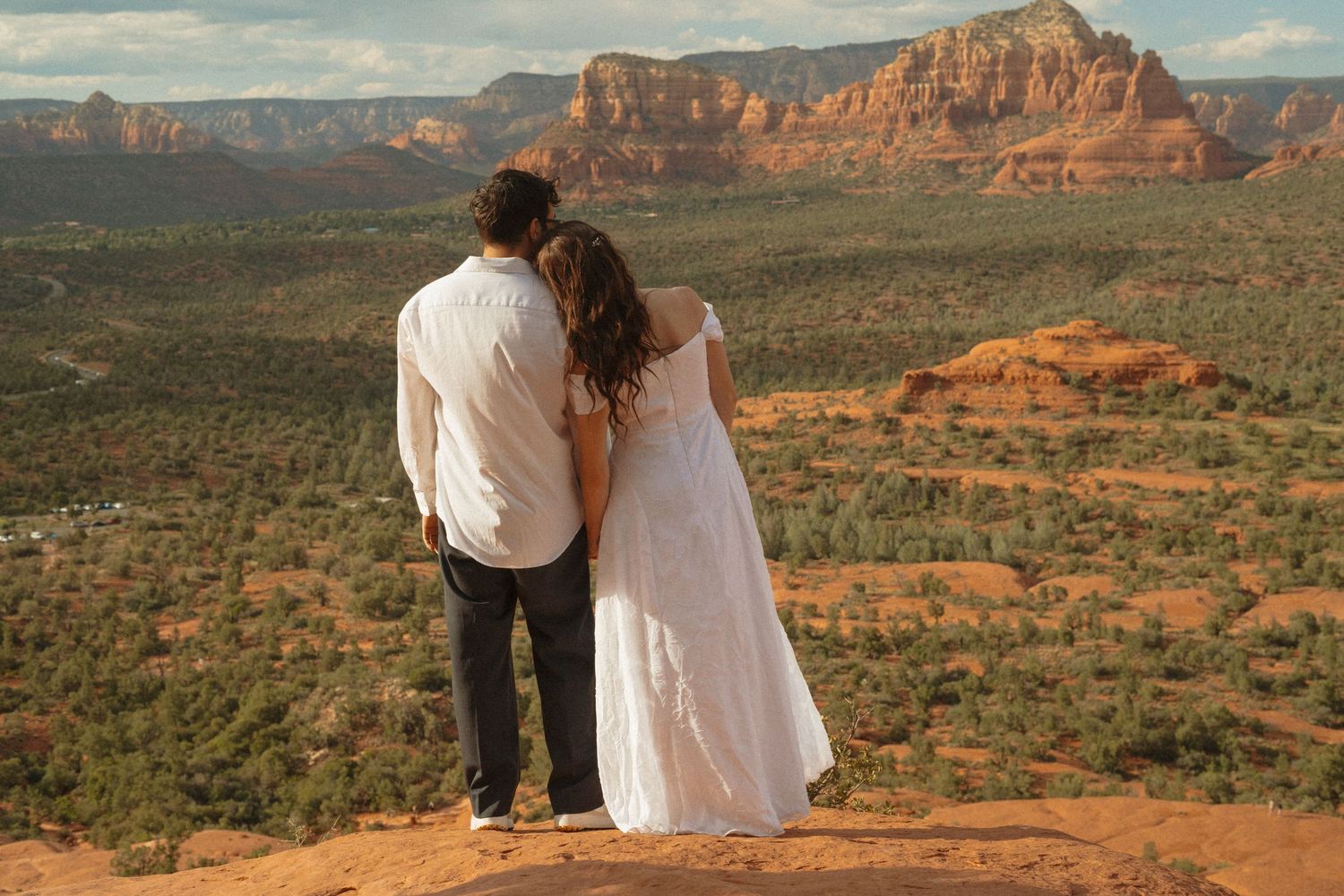 Couple in white dress and dark pants embracing on red rock overlook with Sedona Arizona landscape behind them.