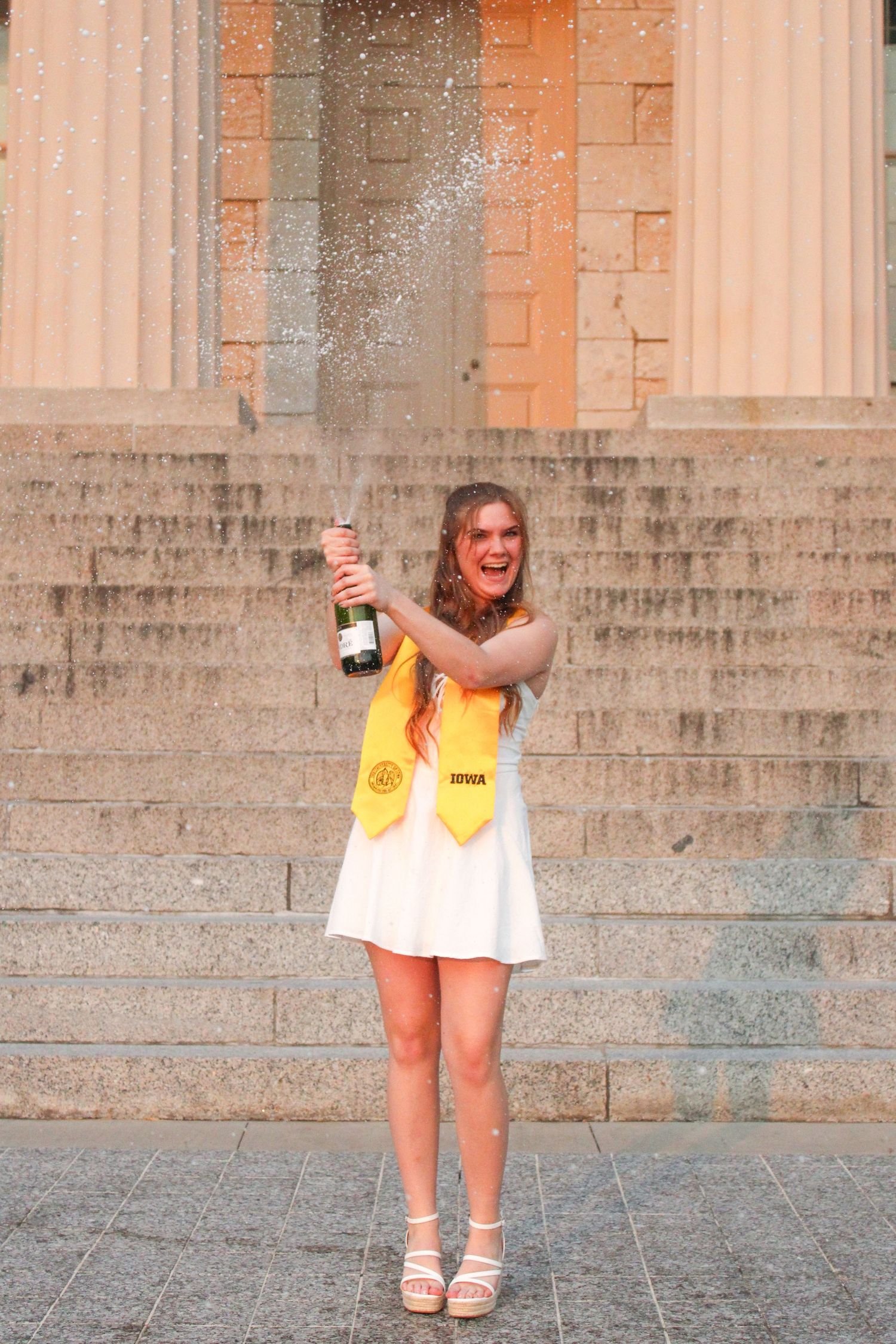Graduate in yellow stole celebrates with champagne spray on stone steps.