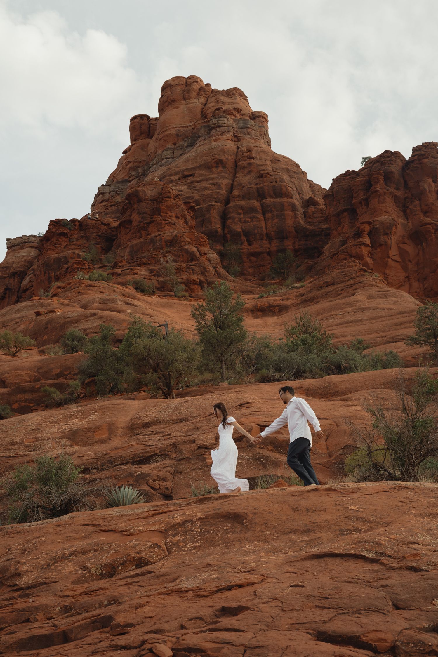 Couple in wedding attire holding hands on red rock formations in Sedona, Arizona, with iconic buttes in the background.