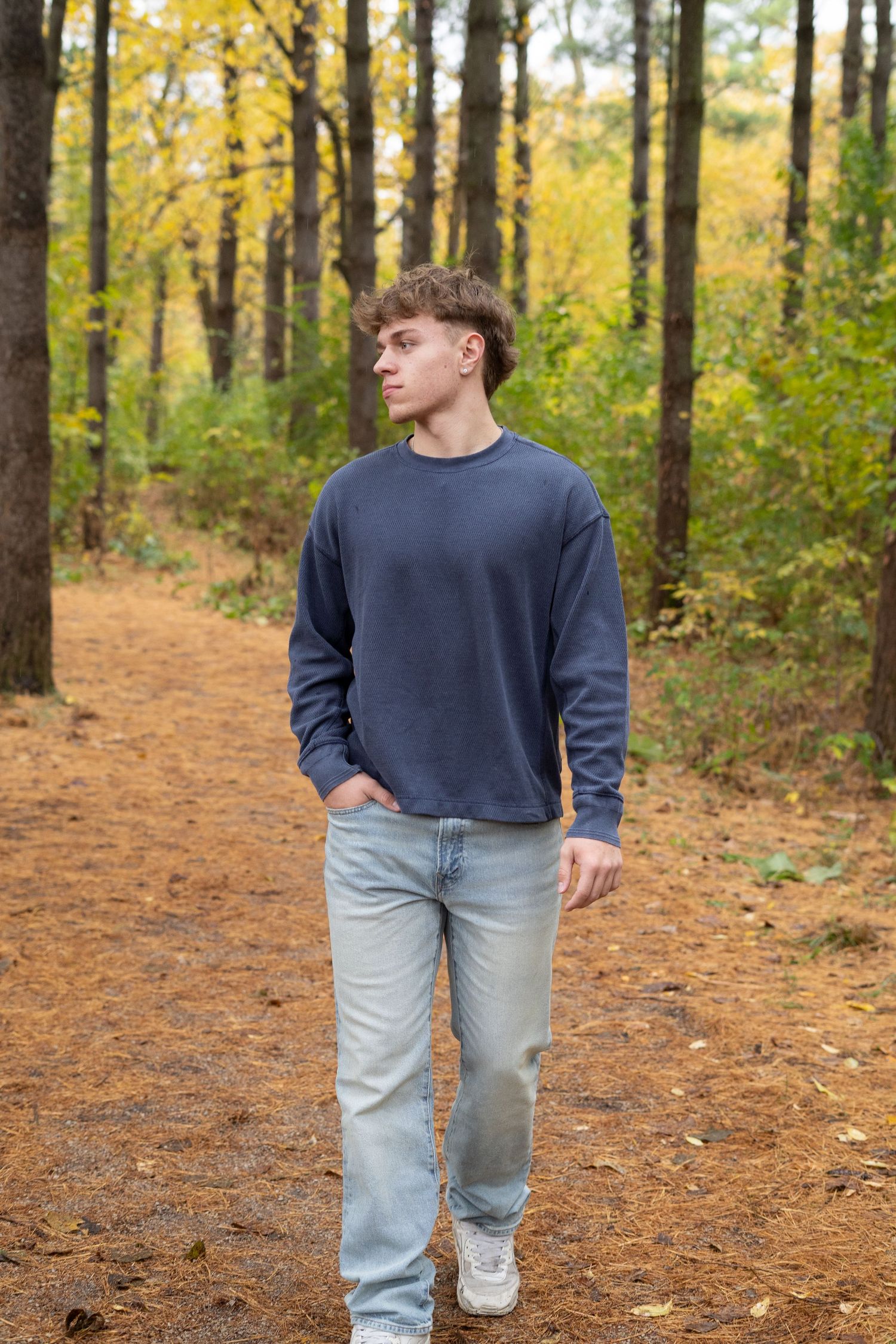 Person wearing navy sweater and light jeans walks along autumn forest trail covered in fallen leaves.