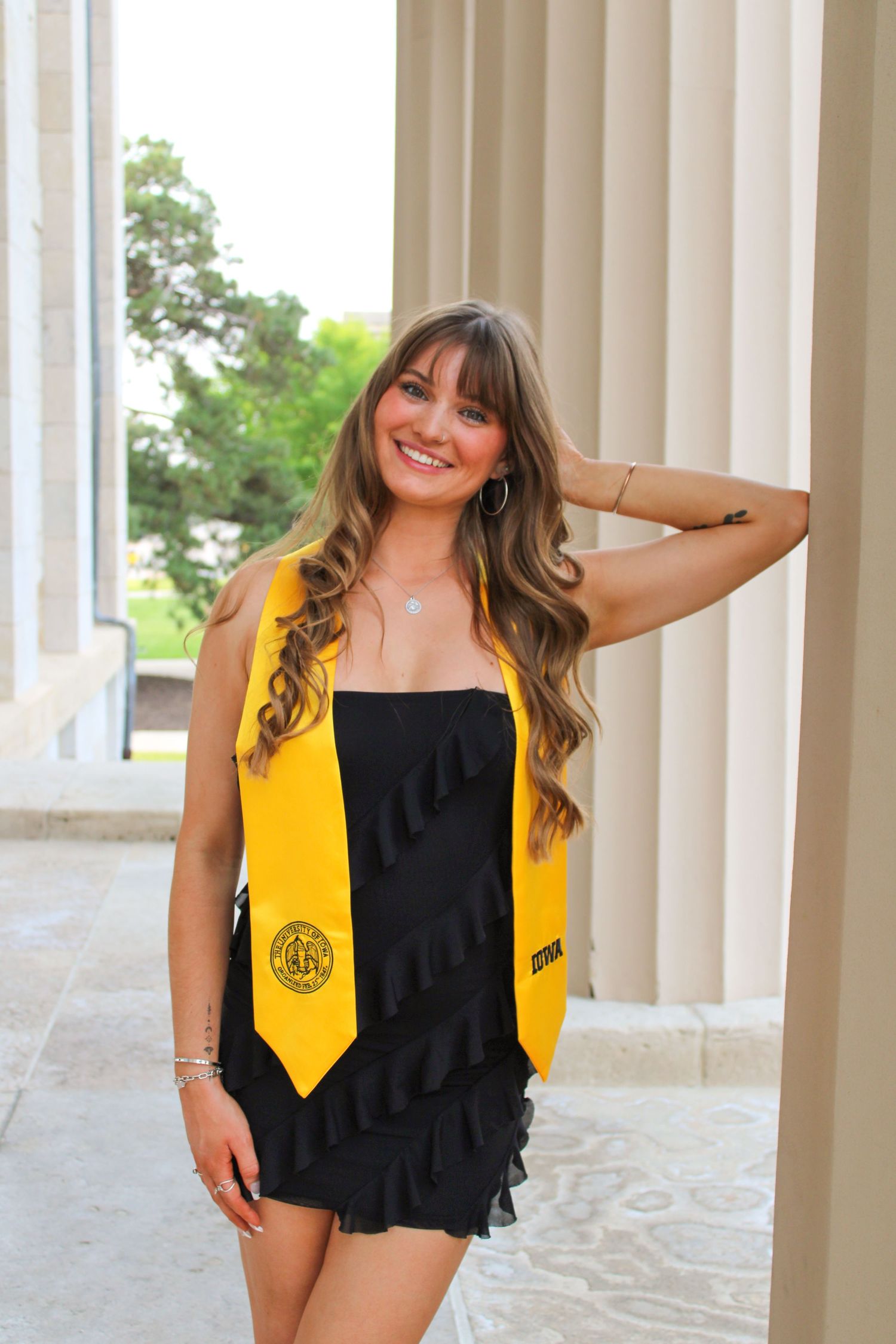 Graduate wearing yellow stole and black dress poses by classical columns during graduation celebration.