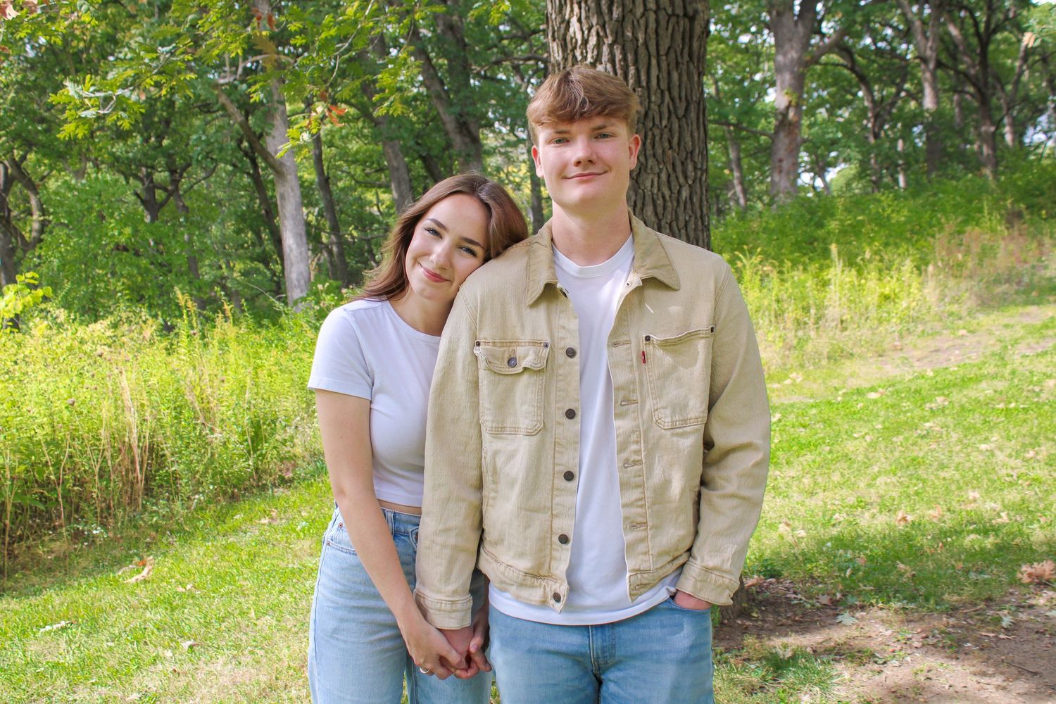 A casual outdoor portrait of a couple standing together near a tree in natural lighting.