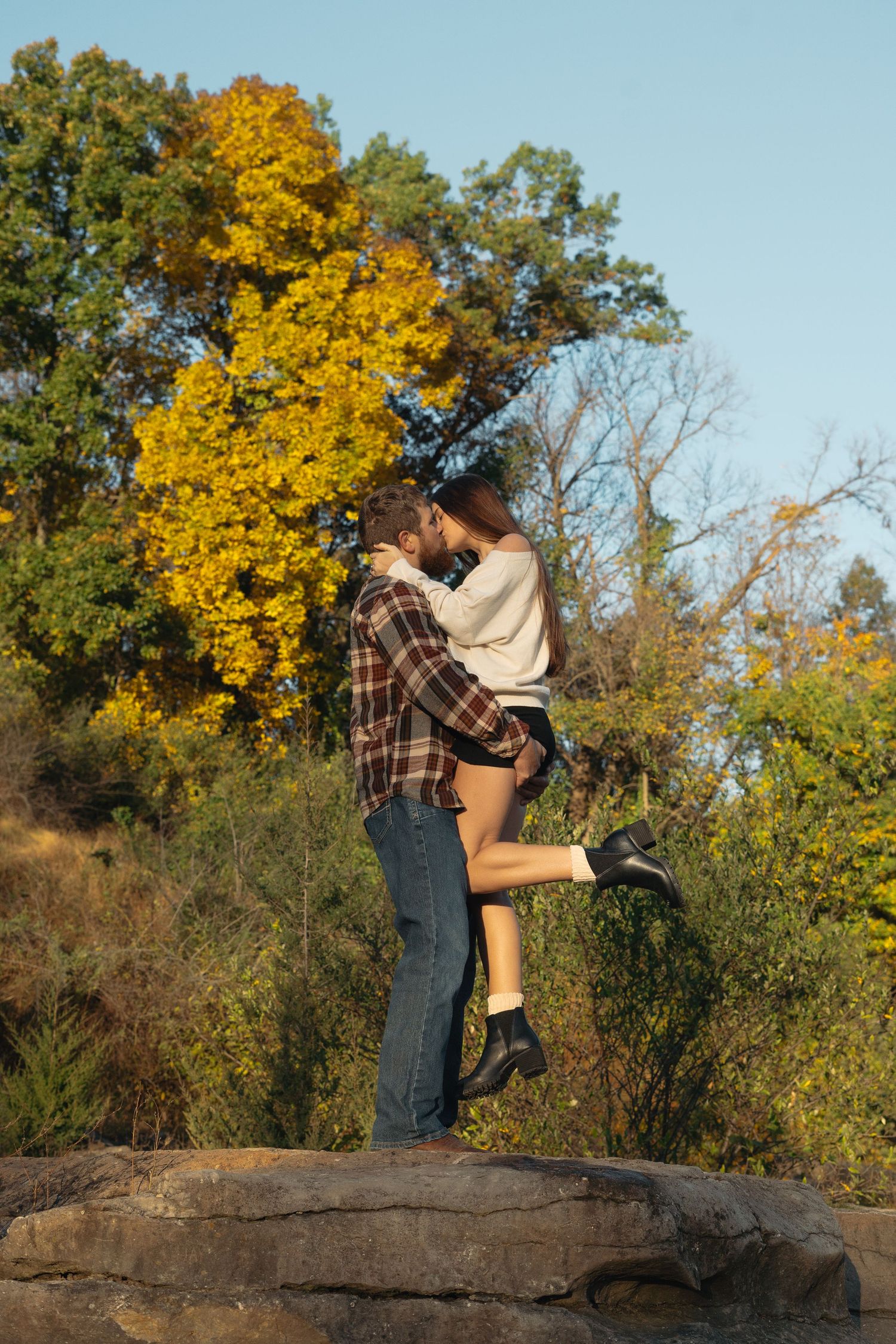 A couple shares a romantic embrace on a rocky overlook during autumn with golden foliage in the background.