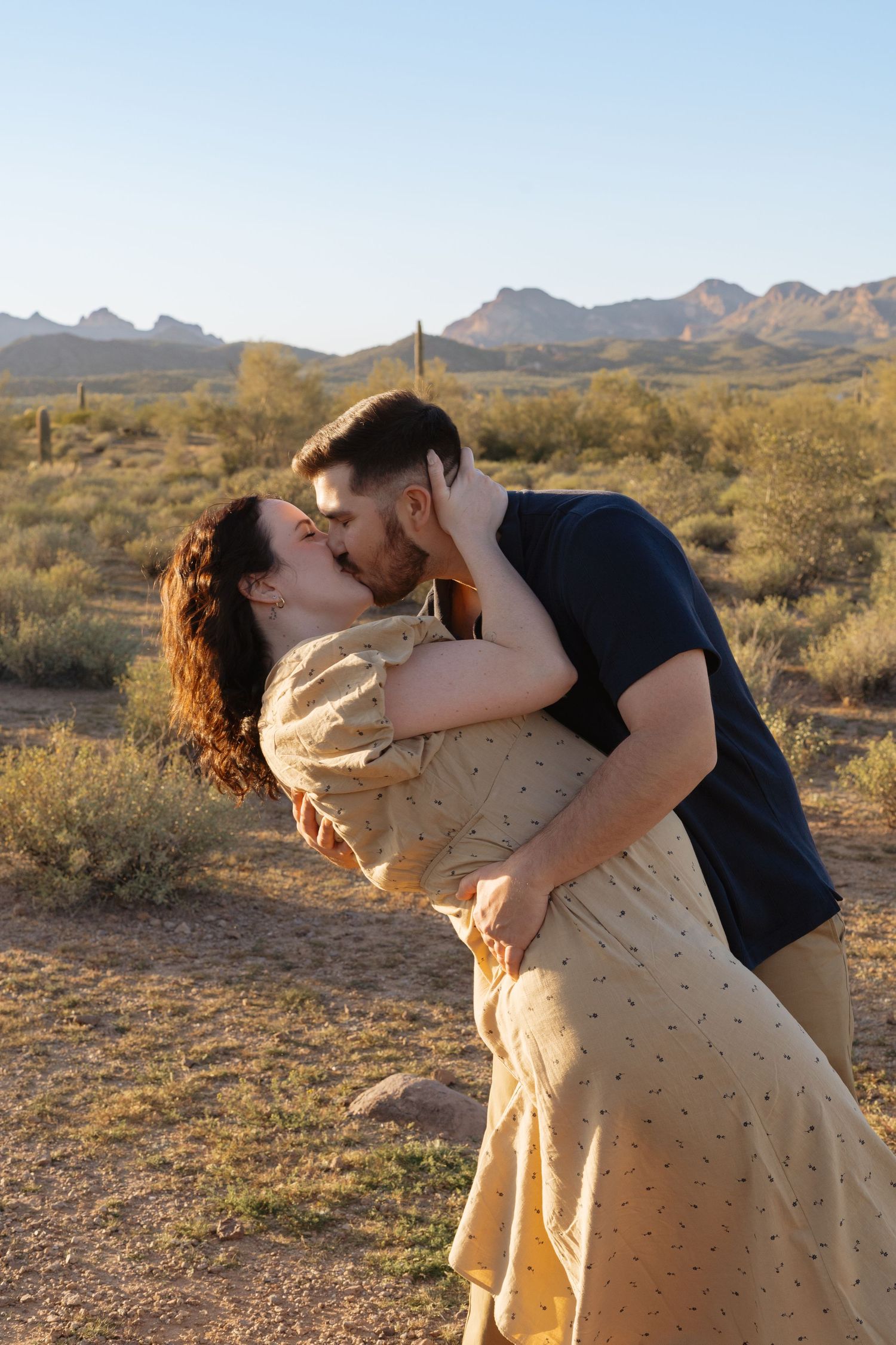 A couple embraces and dips in a romantic pose against a desert mountain landscape at sunset.