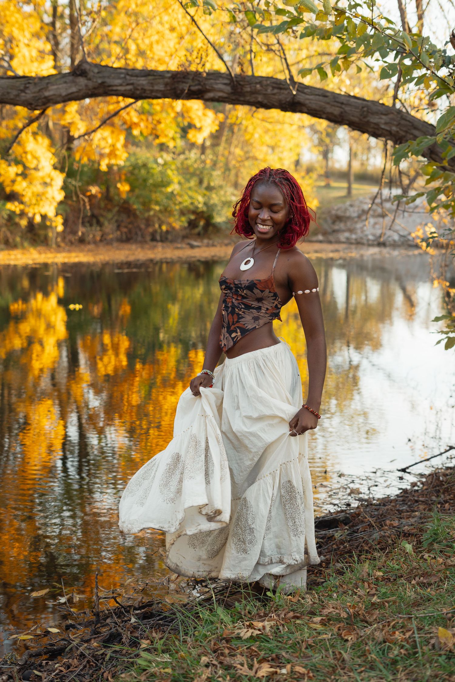 A person in a white dress poses joyfully by a lake surrounded by golden autumn foliage and reflective water.