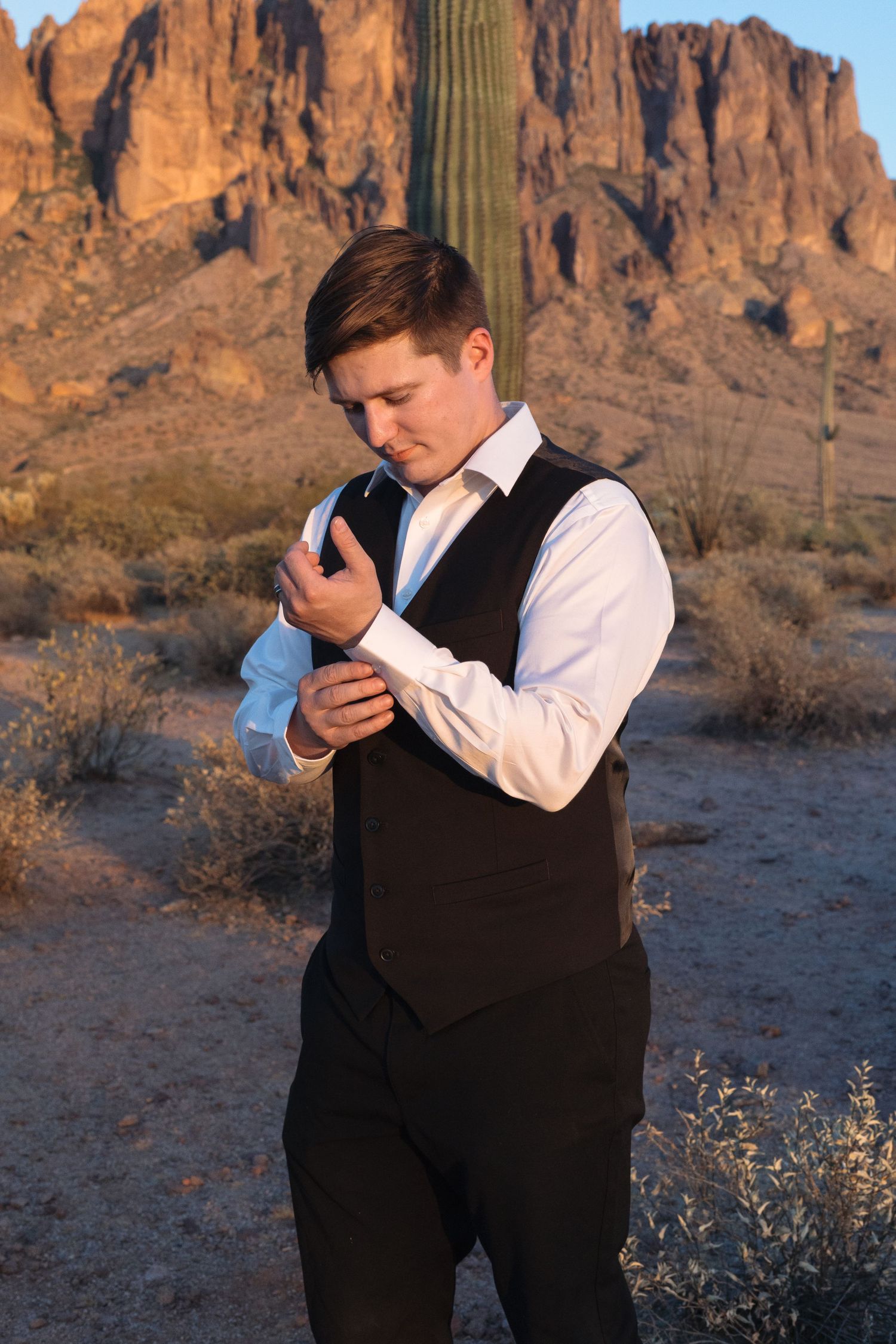 Young man in black vest and white dress shirt adjusting cufflinks in Arizona desert with saguaro cactus and mountains.