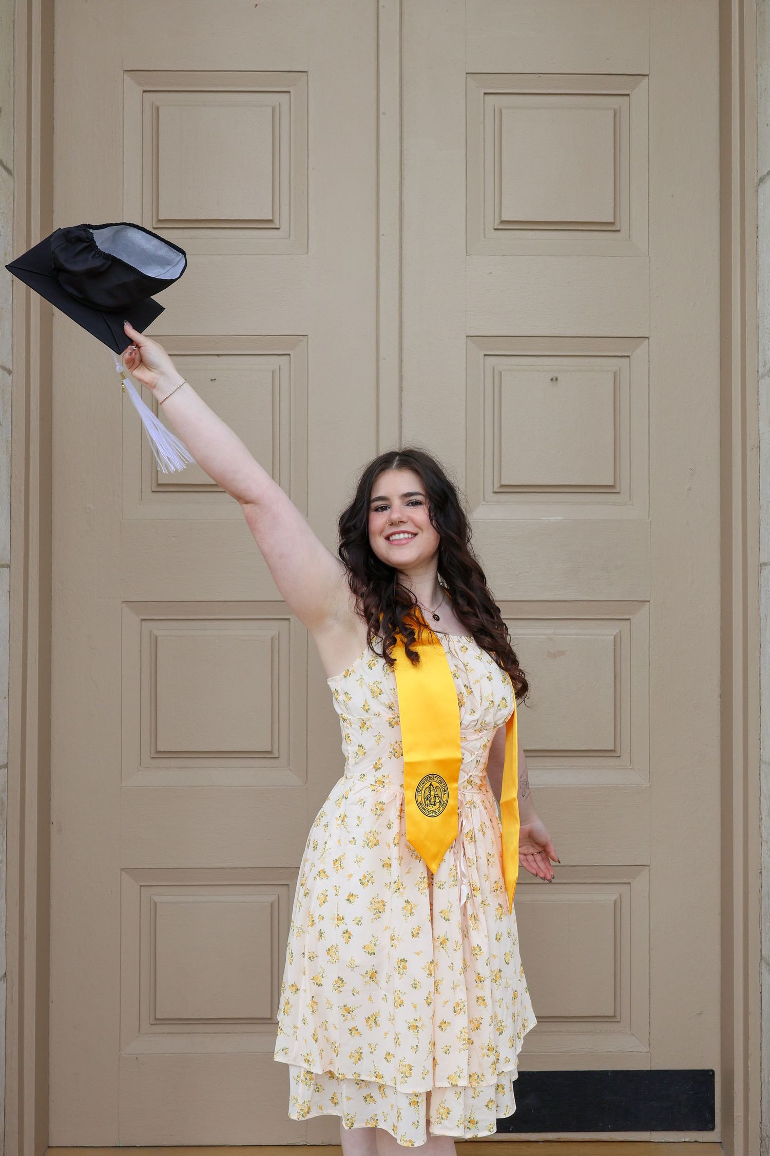 A graduate in a floral dress and yellow sash celebrates with raised arms in front of a beige wall.