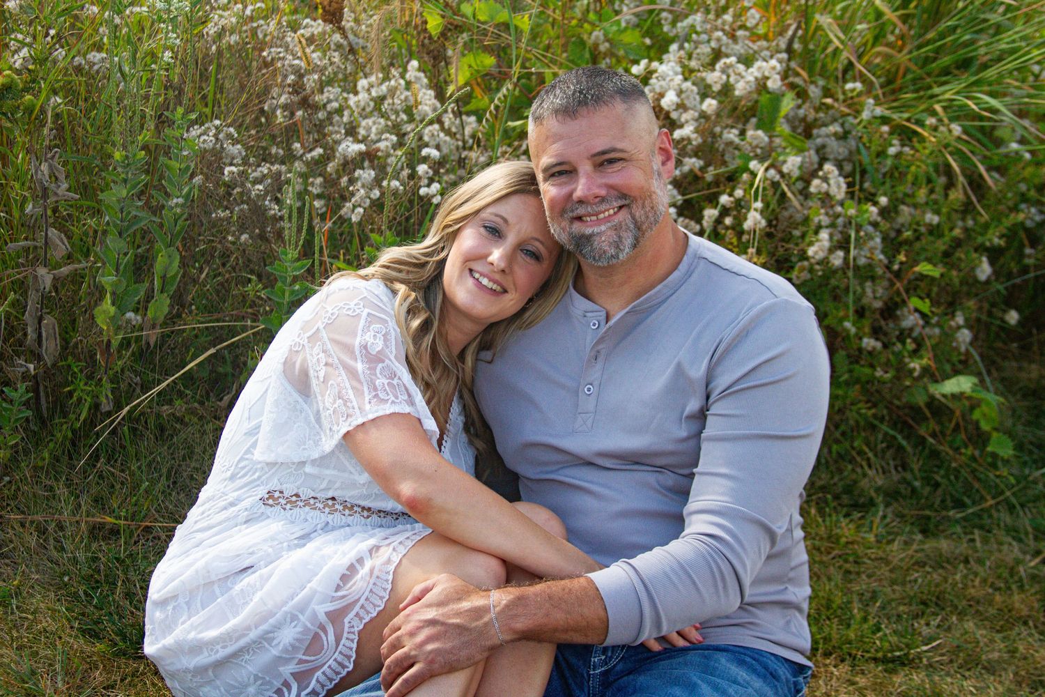 A couple embraces while sitting outdoors among flowering plants and greenery.