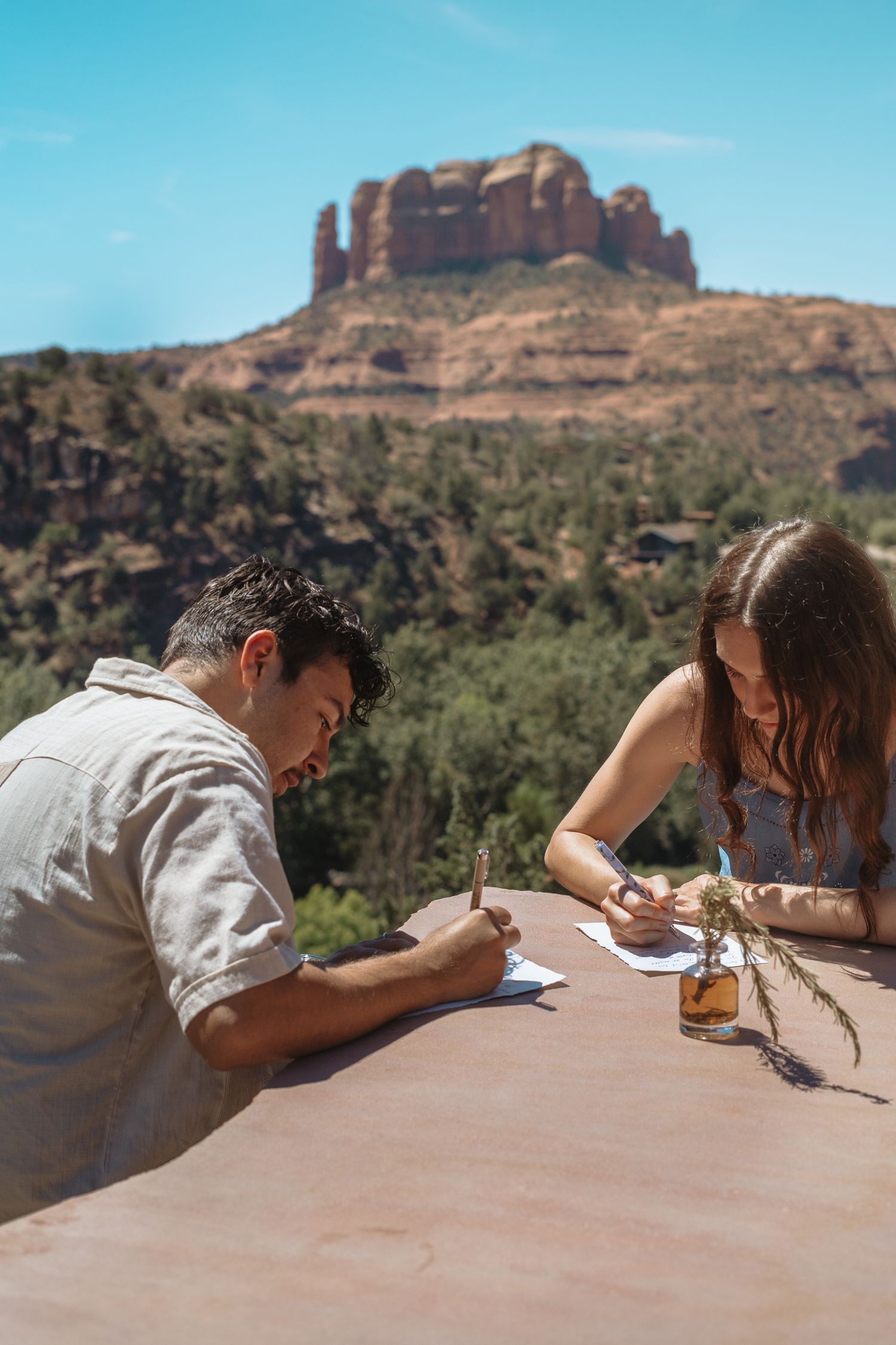 Couple writing vows at outdoor table with Sedona red rock formations and blue sky in background.