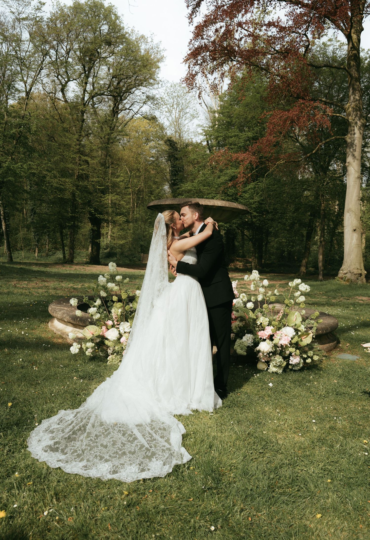 Bride and groom share first kiss at outdoor woodland wedding ceremony with floral arrangements and lace veil.