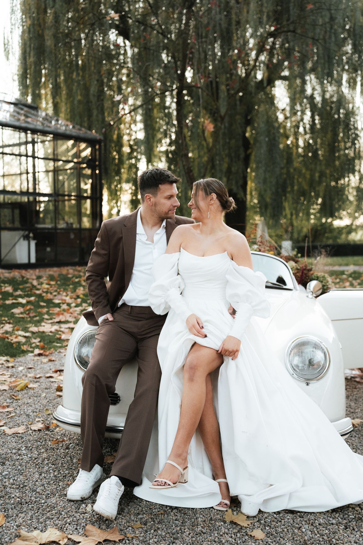 Romantic wedding couple poses together on vintage car surrounded by autumn trees.