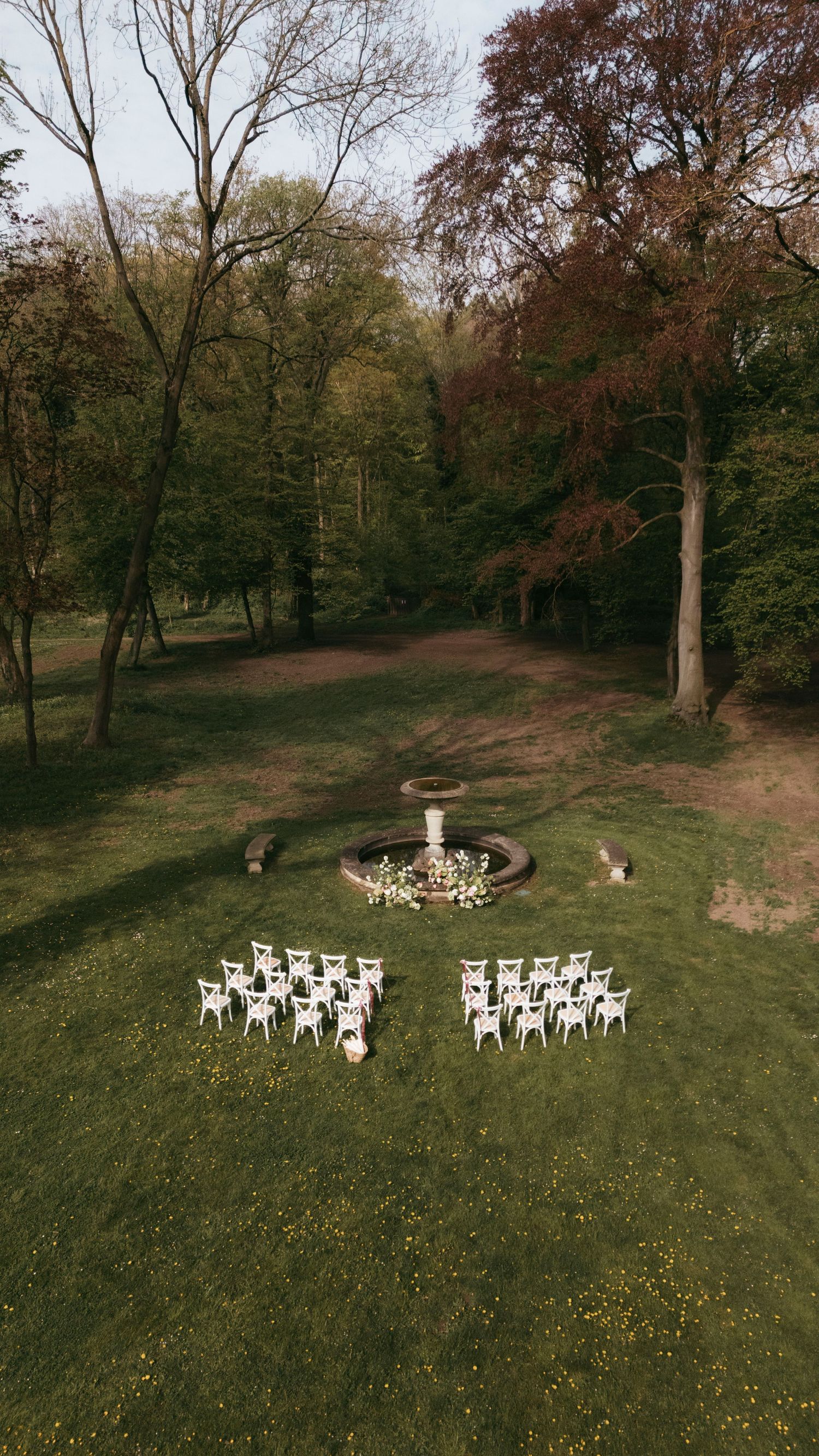 Aerial view of an outdoor wedding ceremony setup with white chairs around a floral fountain in a lush green park.