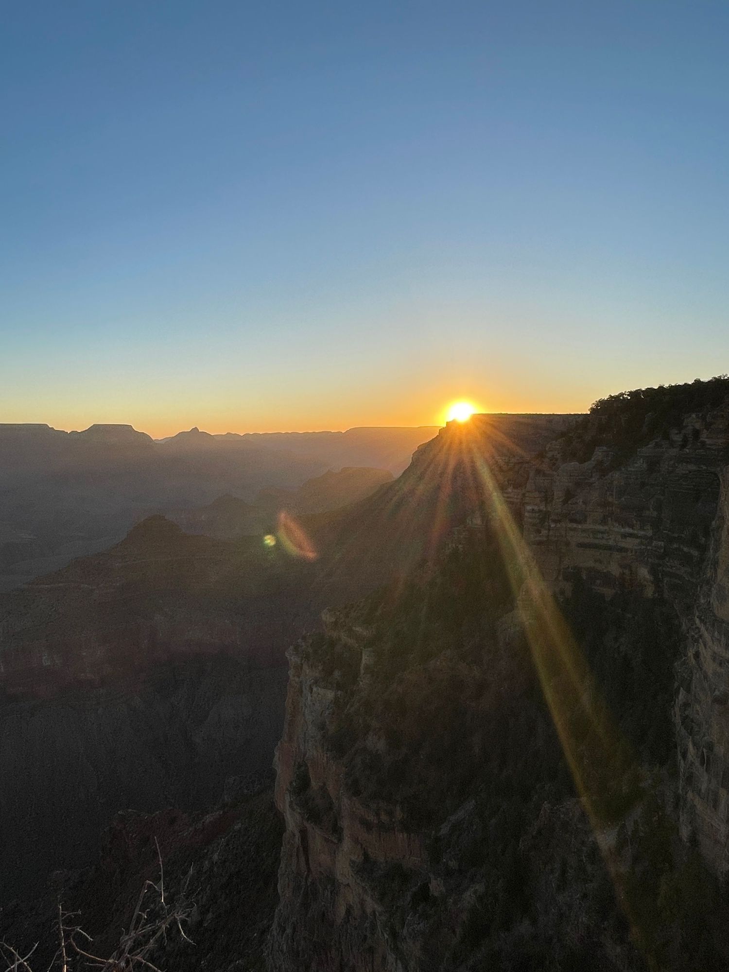 Stunning sunset over the Grand Canyon with golden sun rays illuminating layered rock formations under a clear blue sky.