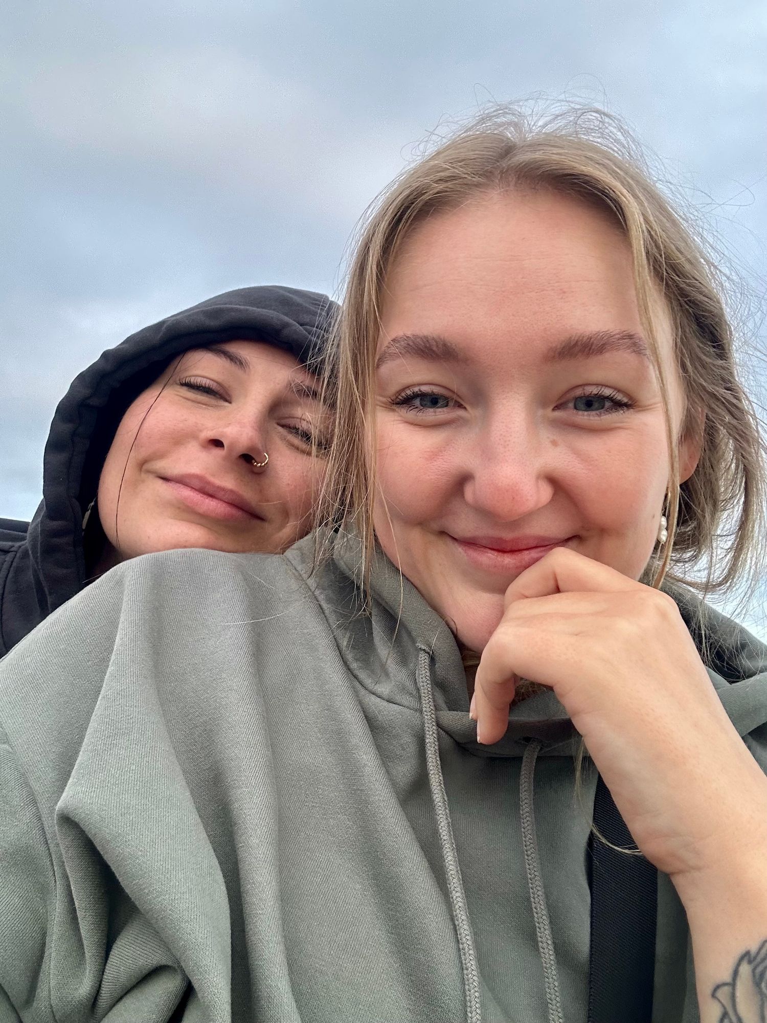 Two young women in hoodies smiling for a selfie outdoors under a cloudy sky.