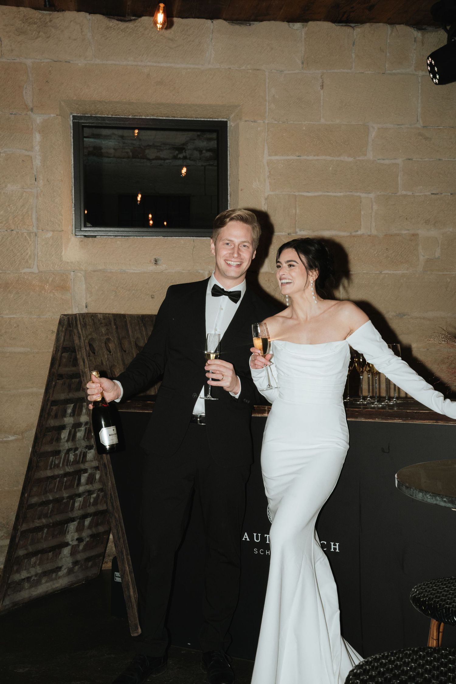 A couple in formal wedding attire share a joyful dance moment in a stone-walled room with dim lighting.