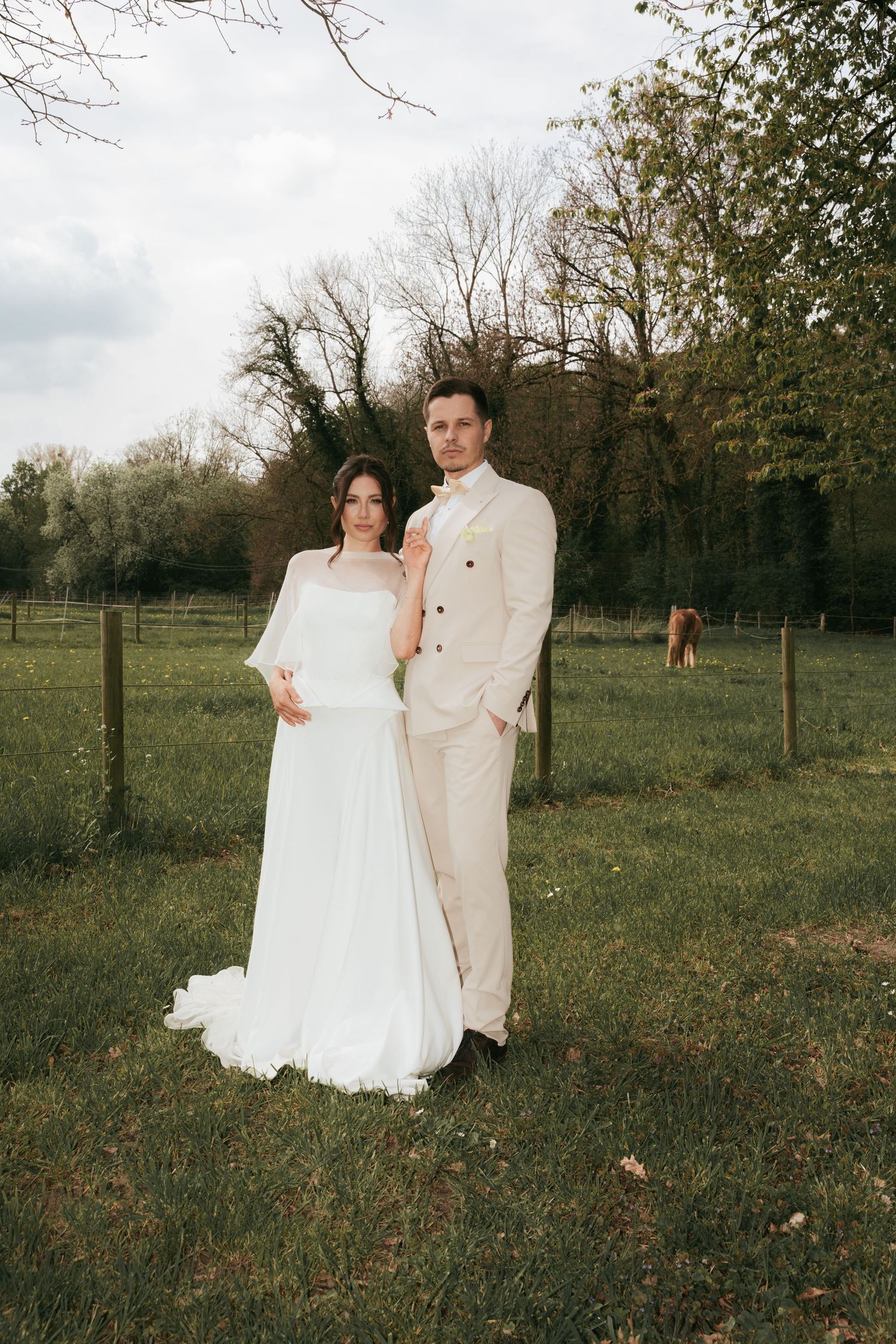 Bride in white gown and groom in beige suit posing together in a green field with trees in background.