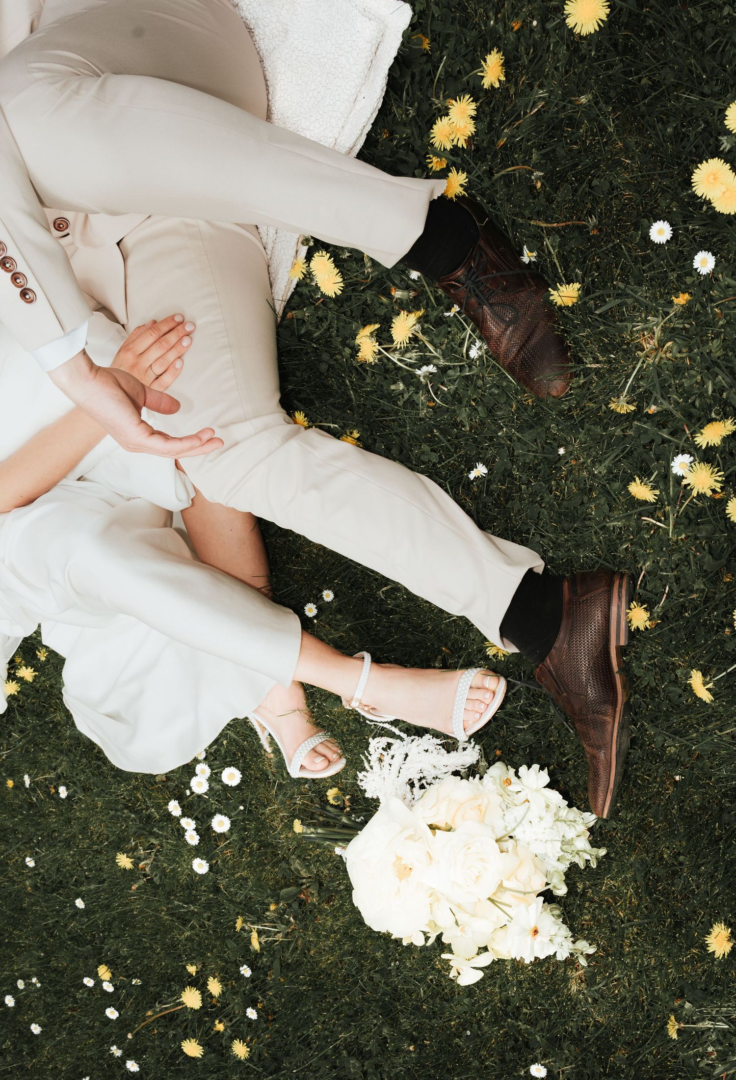 Couple sitting on grass with white bouquet, dandelions, wearing beige outfit and brown shoes.
